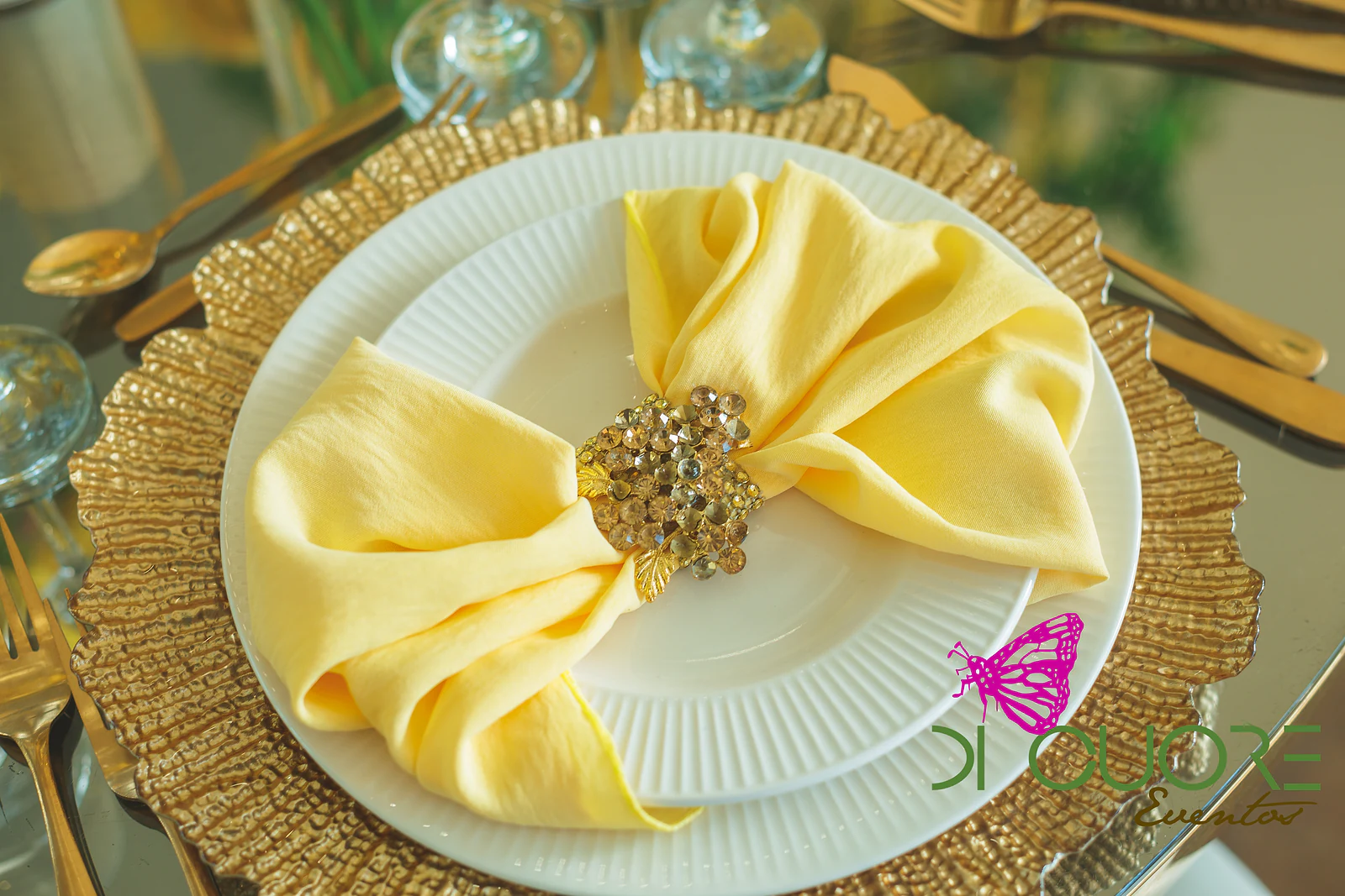 White plate with a pale yellow napkin held by a brooch, on a gold charger with a scalloped edge