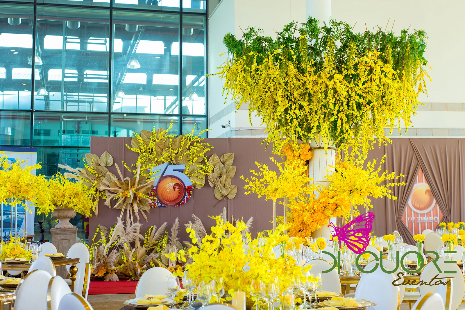 Dusty-pink draped panel with the TV Zimbo logo and the number 15, among dried palm leaves and yellow flowers, beneath a foliage chandelier hanging from a column