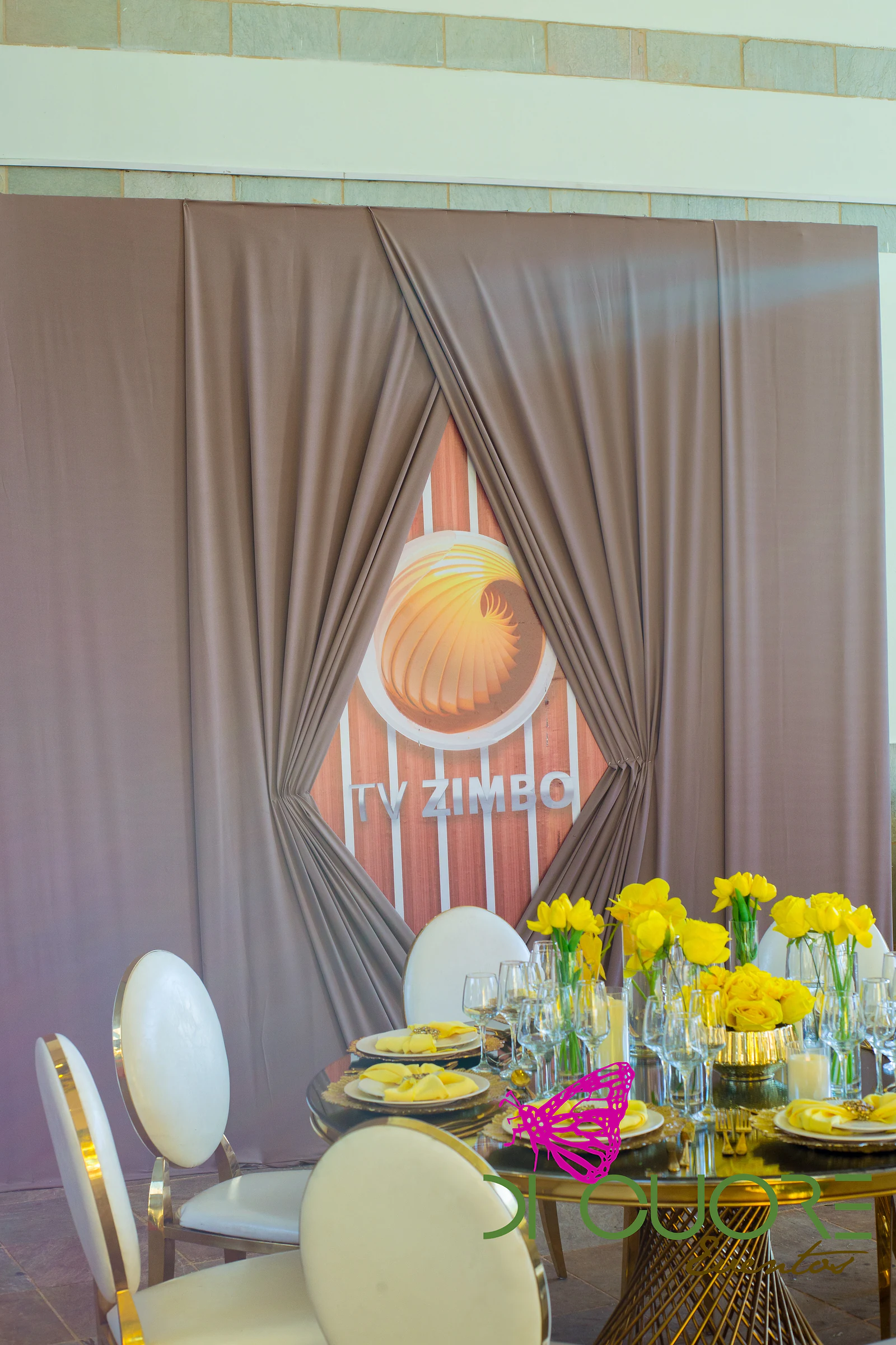 Dusty-pink draped panel with the TV Zimbo logo in a diamond and, in front, a round table with yellow tulips and round-backed chairs