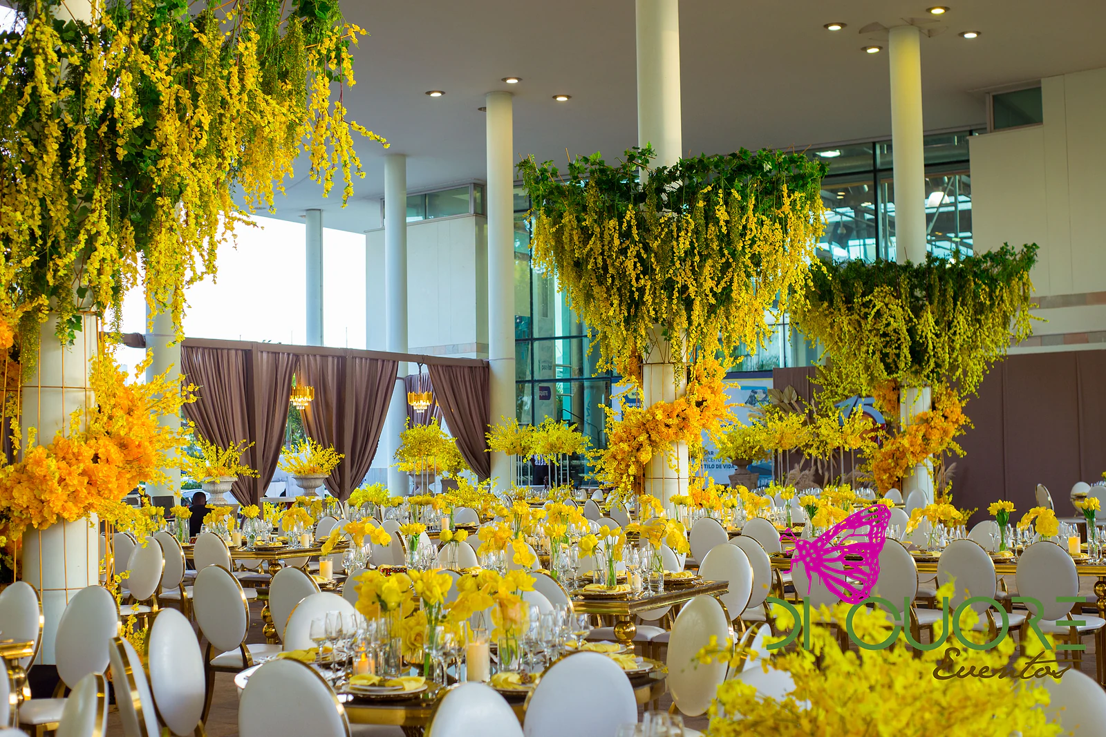 Hall with three chandeliers of foliage and yellow orchids hanging from white columns, above rows of laid tables and round-backed chairs