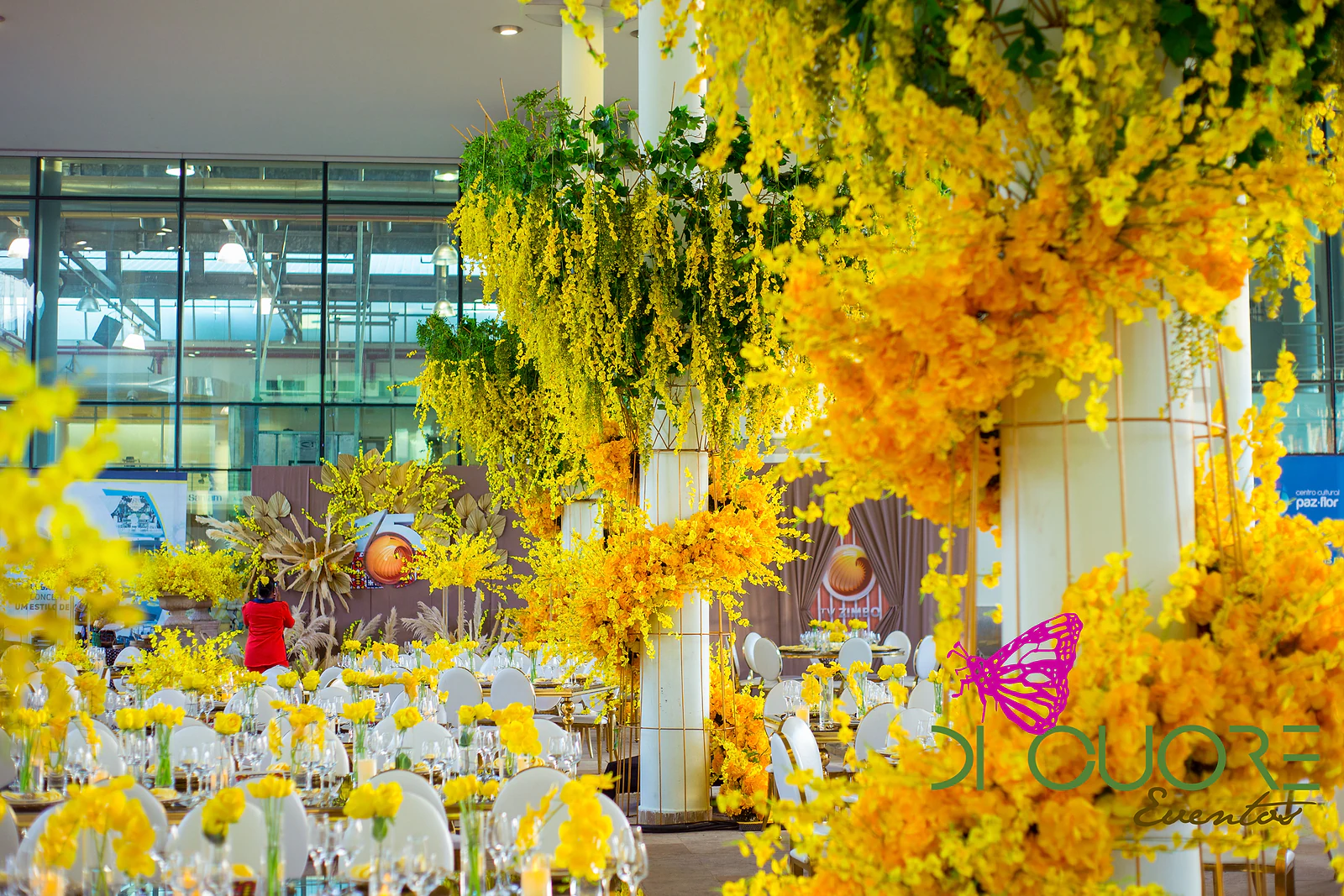 Salão de paredes de vidro iluminado pela luz do dia, com colunas brancas envolvidas por grandes cascatas de flores amarelas e mesas postas ao fundo