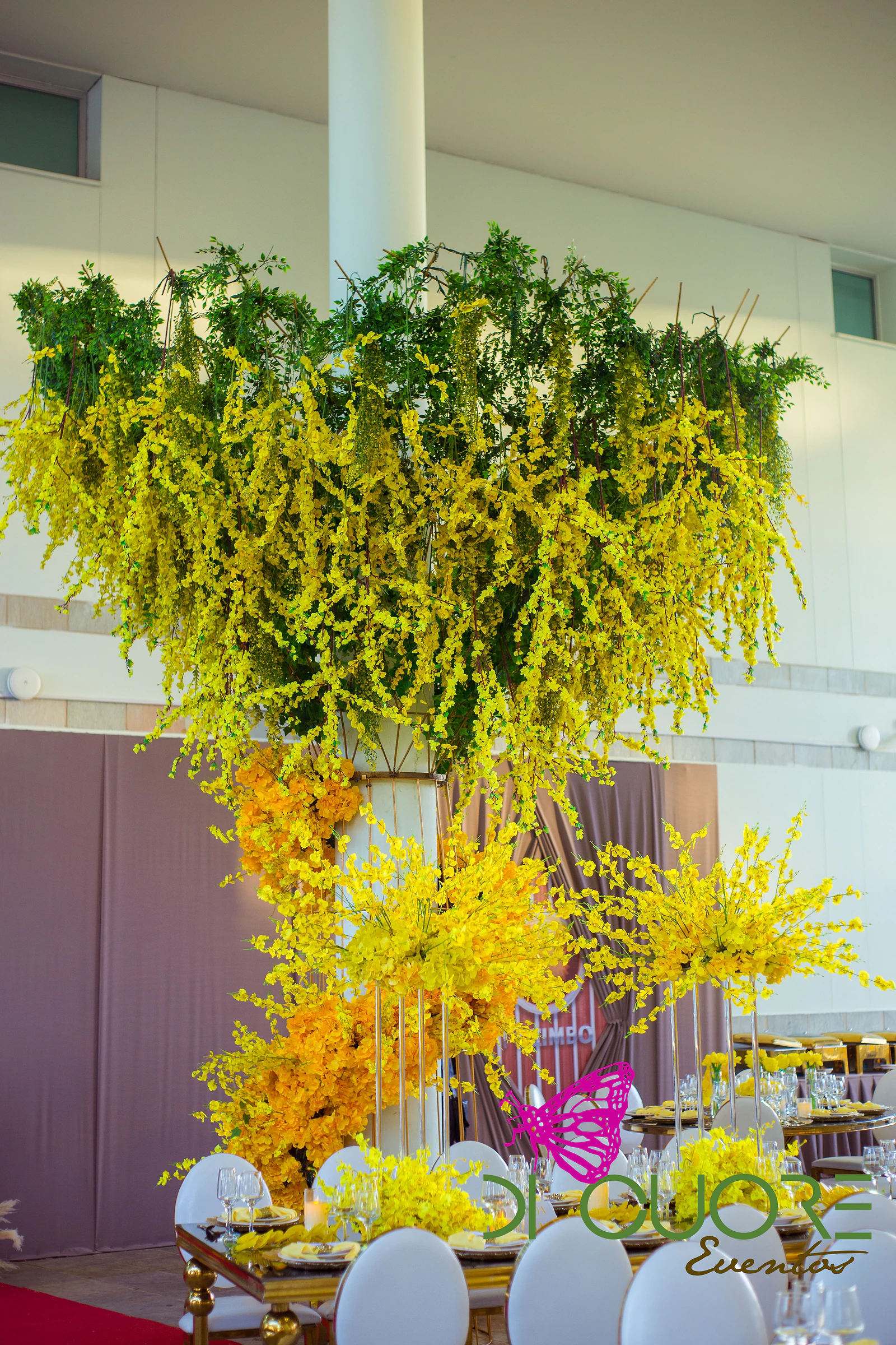 Chandelier of foliage and yellow orchids hanging from a white column, with orange orchids at its base and laid tables with round-backed chairs
