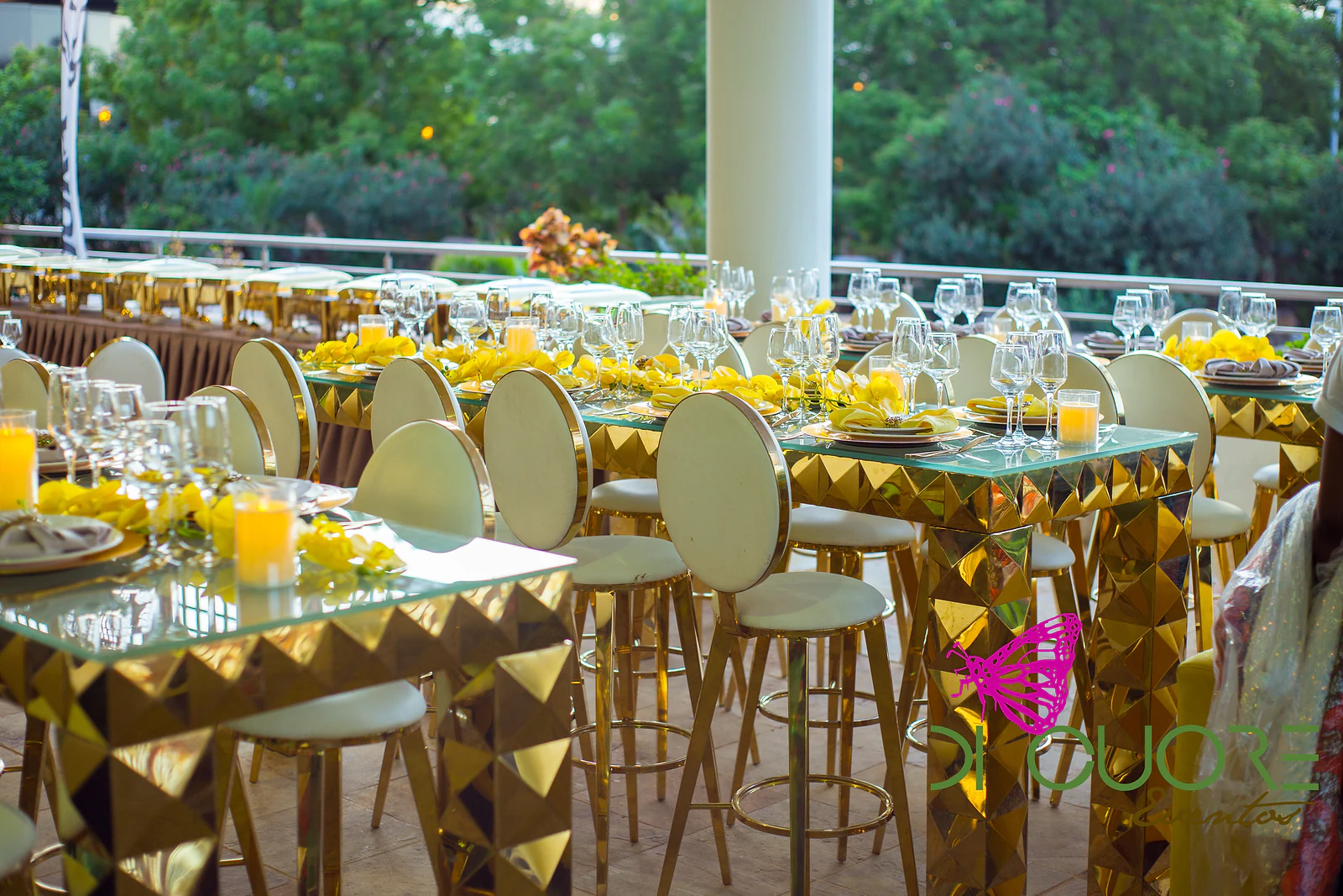 Tall table of gold pyramids with a glass top, candles and high stools with round white backs, facing the garden