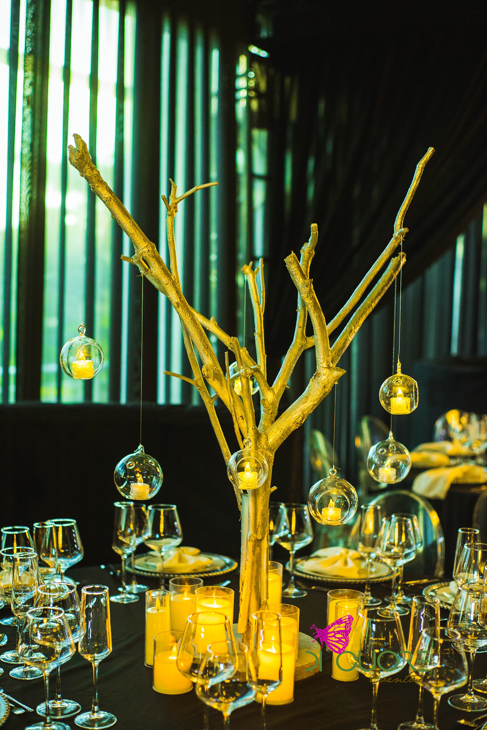 Ramo dourado com globos de vidro e velas, rodeado de copos de cristal e velas de várias alturas, diante de cortinados às riscas