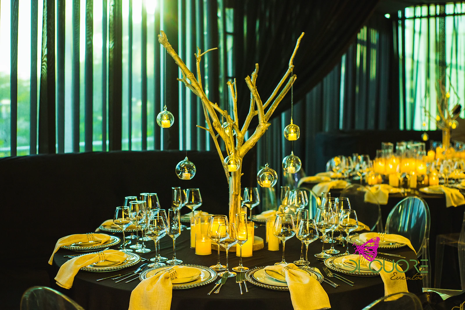 Round table with a black cloth, a tree of golden branches, candles and hanging glass spheres, set against dark drapes