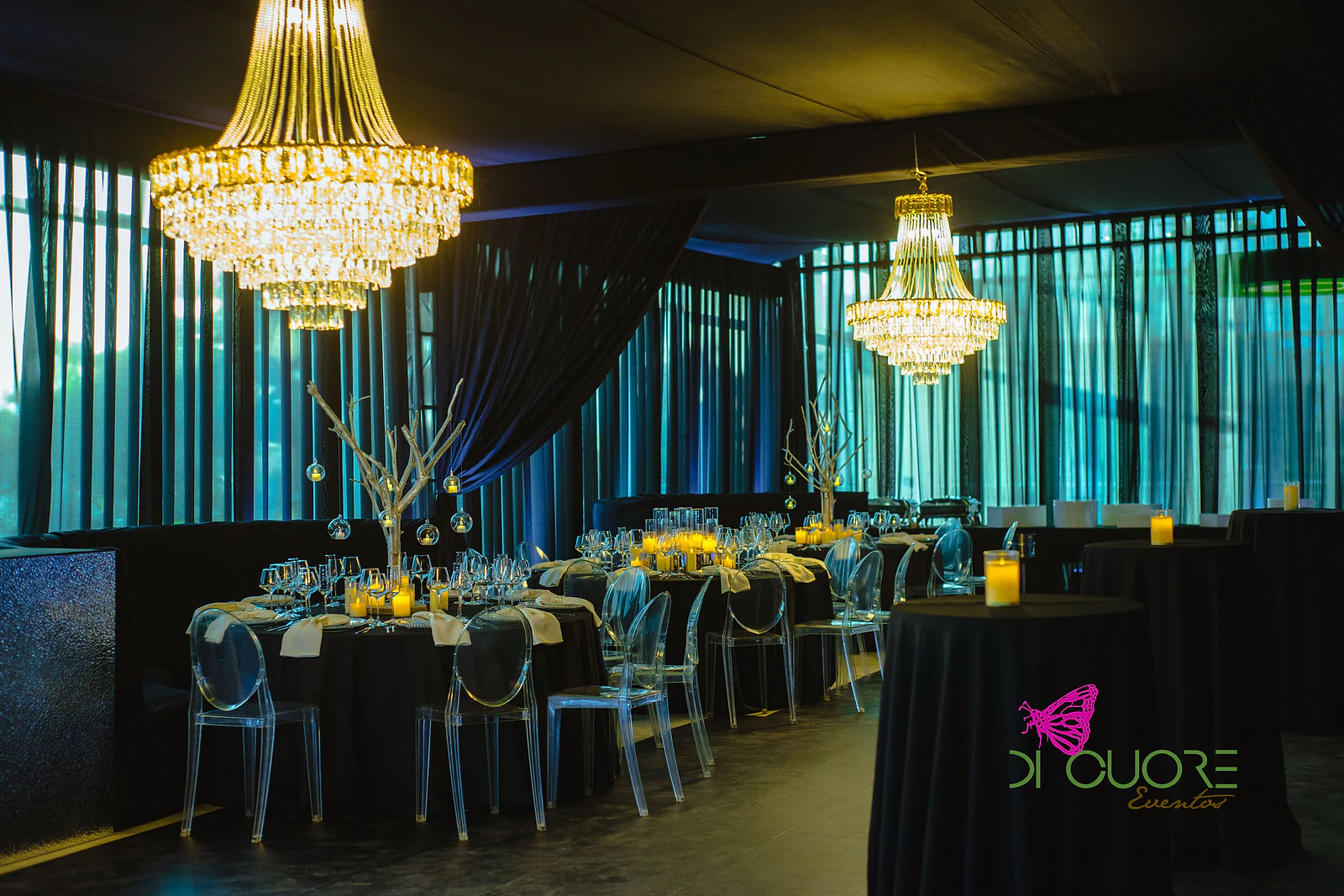 Sala com dois lustres de cristal, mesas redondas de toalha negra com cadeiras transparentes e cortinados às riscas com luz verde-azulada
