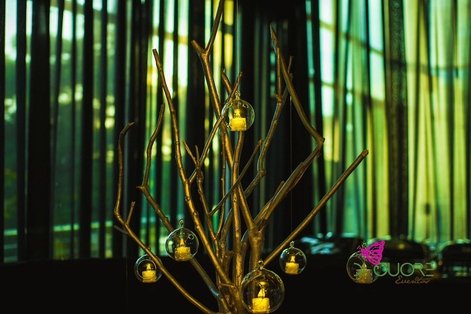 Ramo dourado com lanternas de vidro suspensas e velas acesas, diante de cortinados às riscas com luz verde