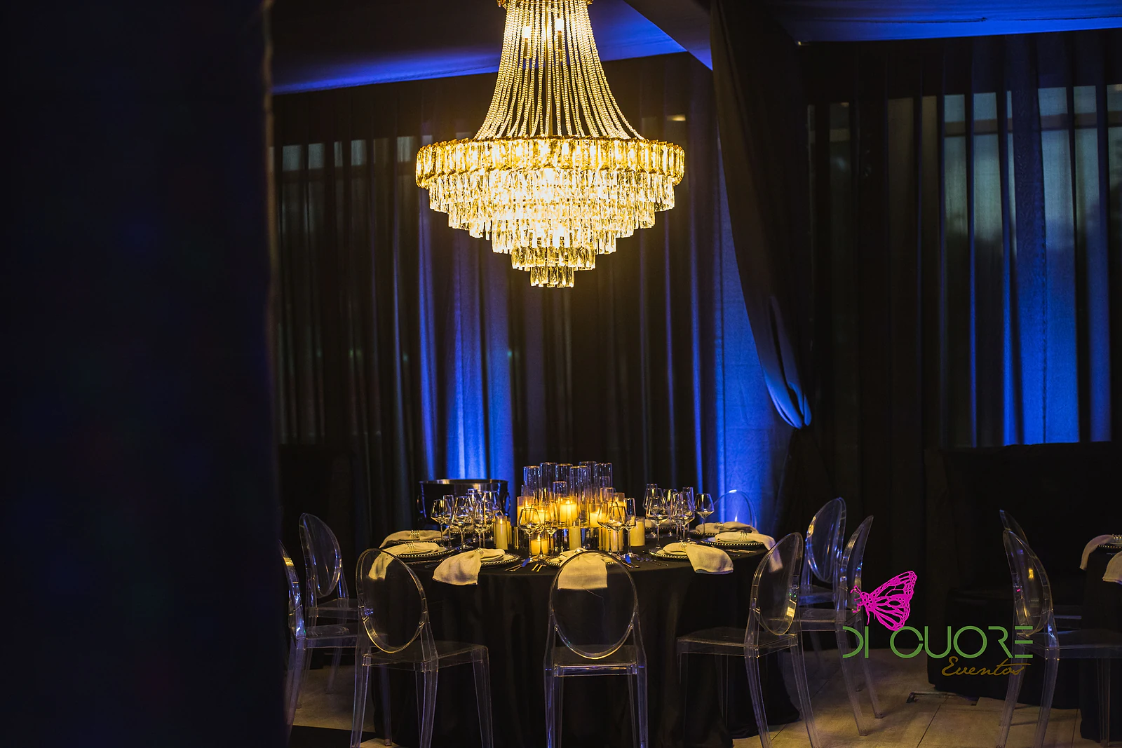 Mesa redonda de toalha negra com cadeiras transparentes e velas, sob um lustre de cristal e cortinados com luz azul