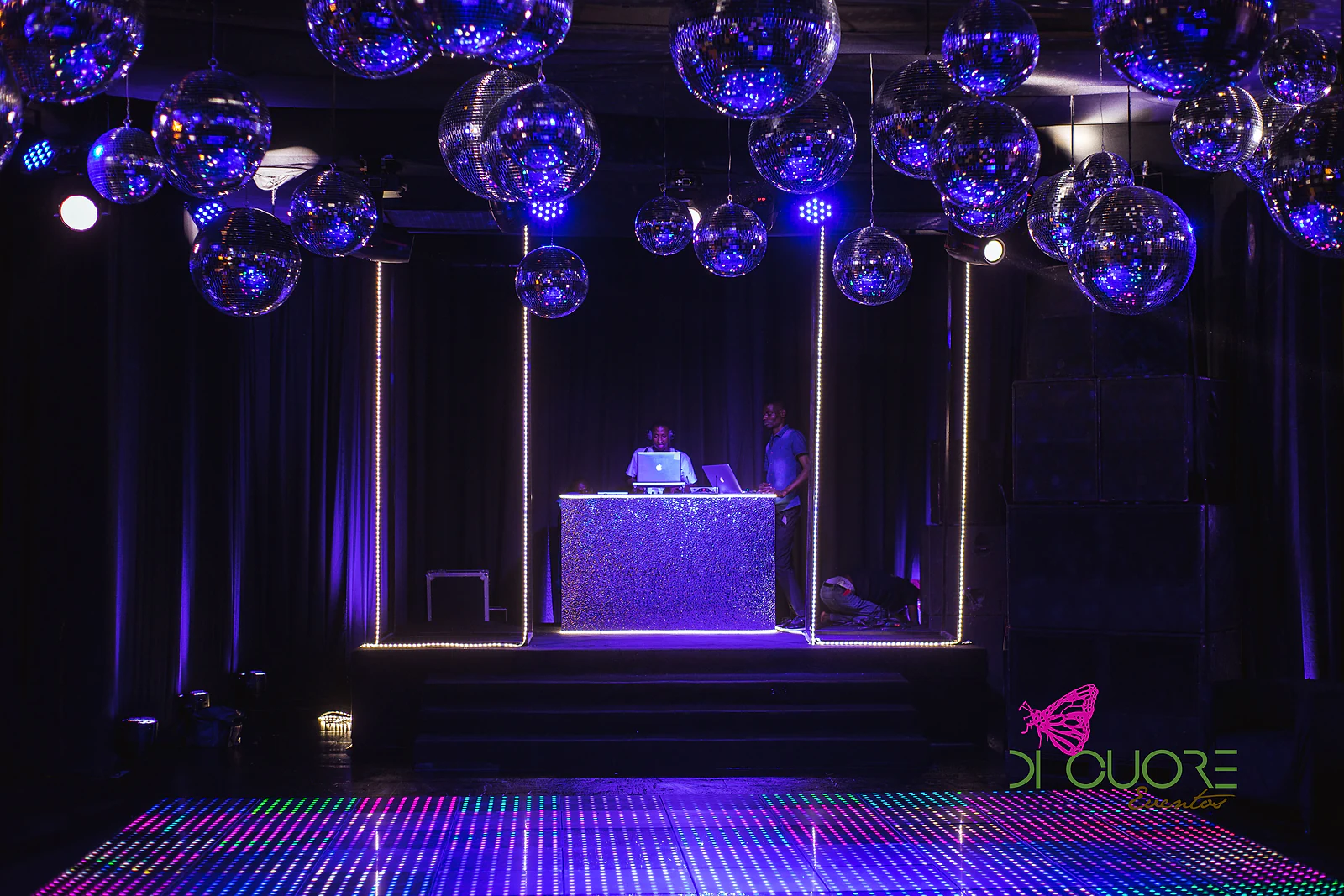 Mesa do DJ com frente brilhante e moldura de fios de luz, esferas de espelho suspensas e pista de dança luminosa em azul e violeta