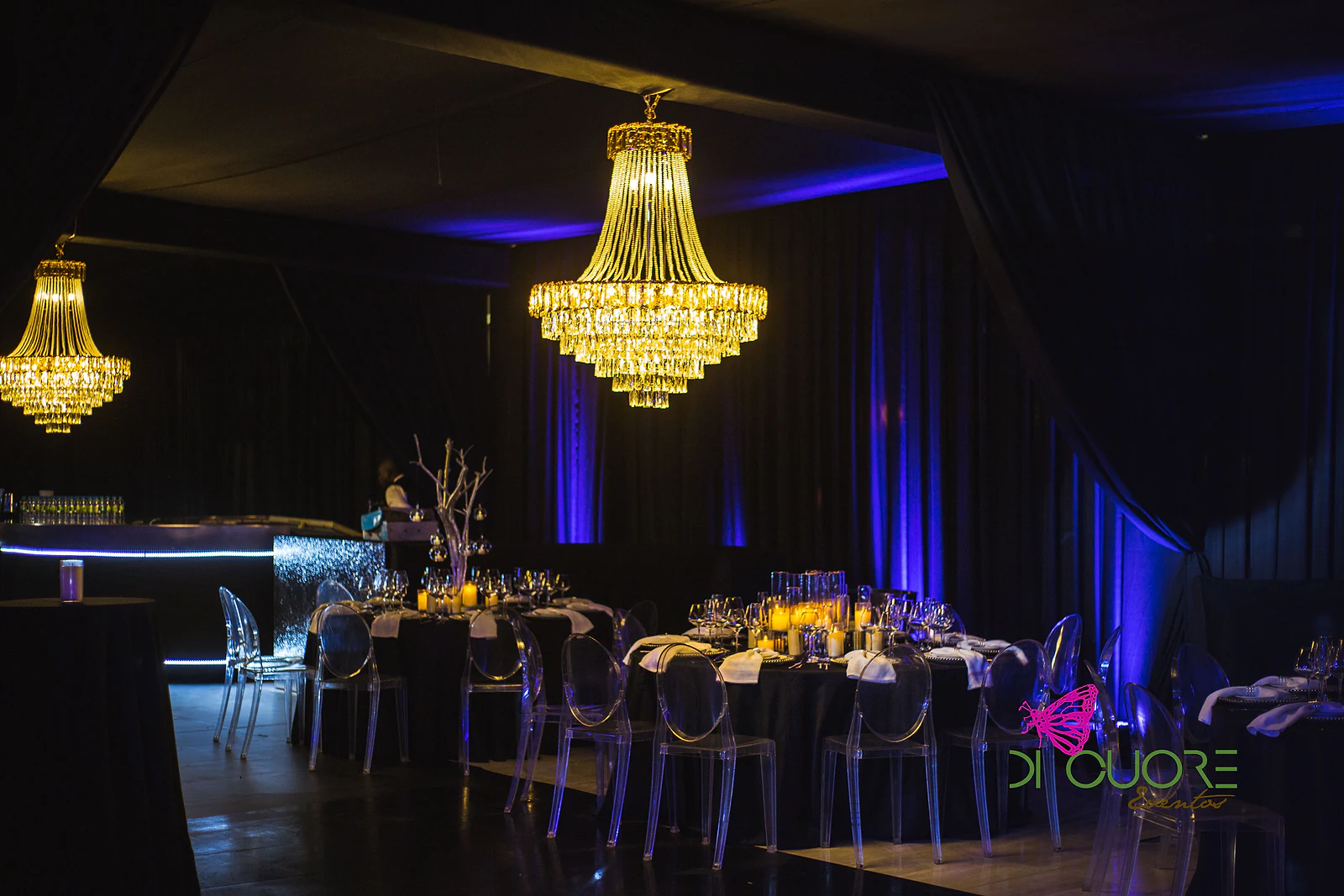 Sala de cortinados negros com lustres de cristal, mesas postas com cadeiras transparentes e um balcão de bar iluminado, sob luz azul