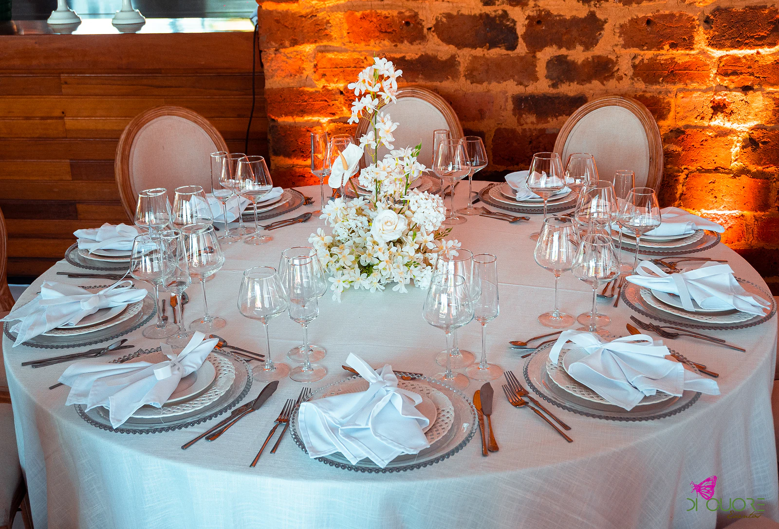 Round table in a pale cloth with ribbon-tied napkins, crystal glasses and a tall spray of white flowers at the centre, before a brick wall