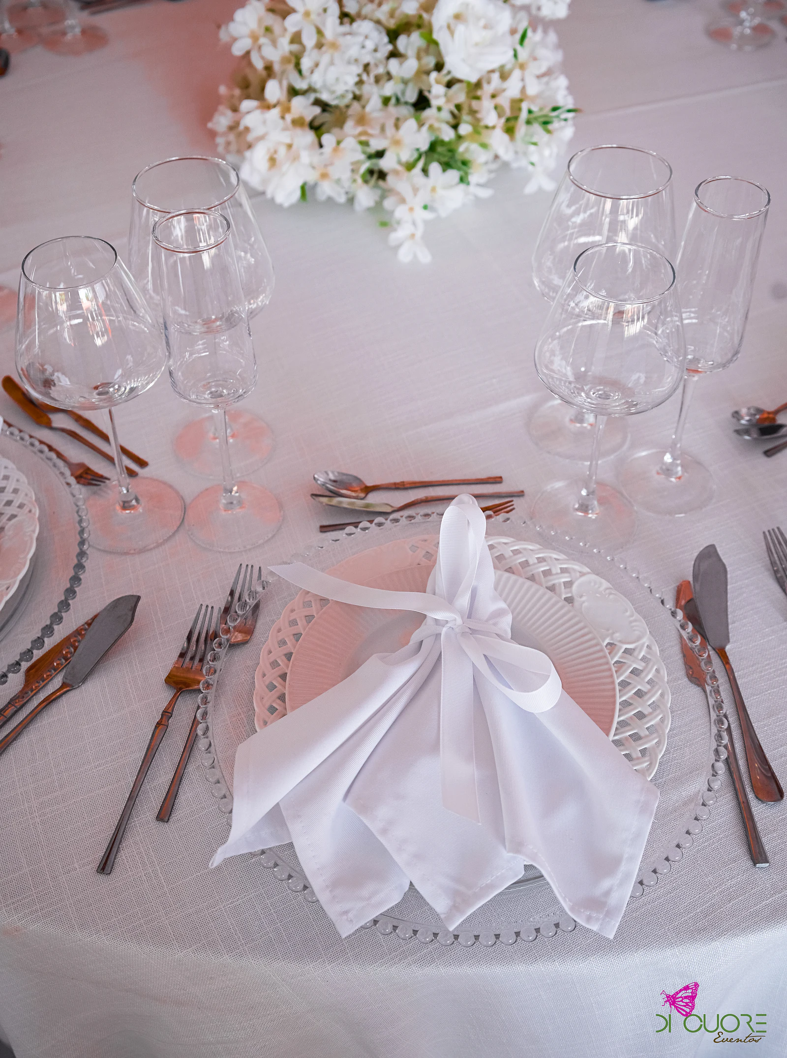 White napkin tied with a ribbon on a beaded glass charger, between crystal glasses and copper-coloured cutlery
