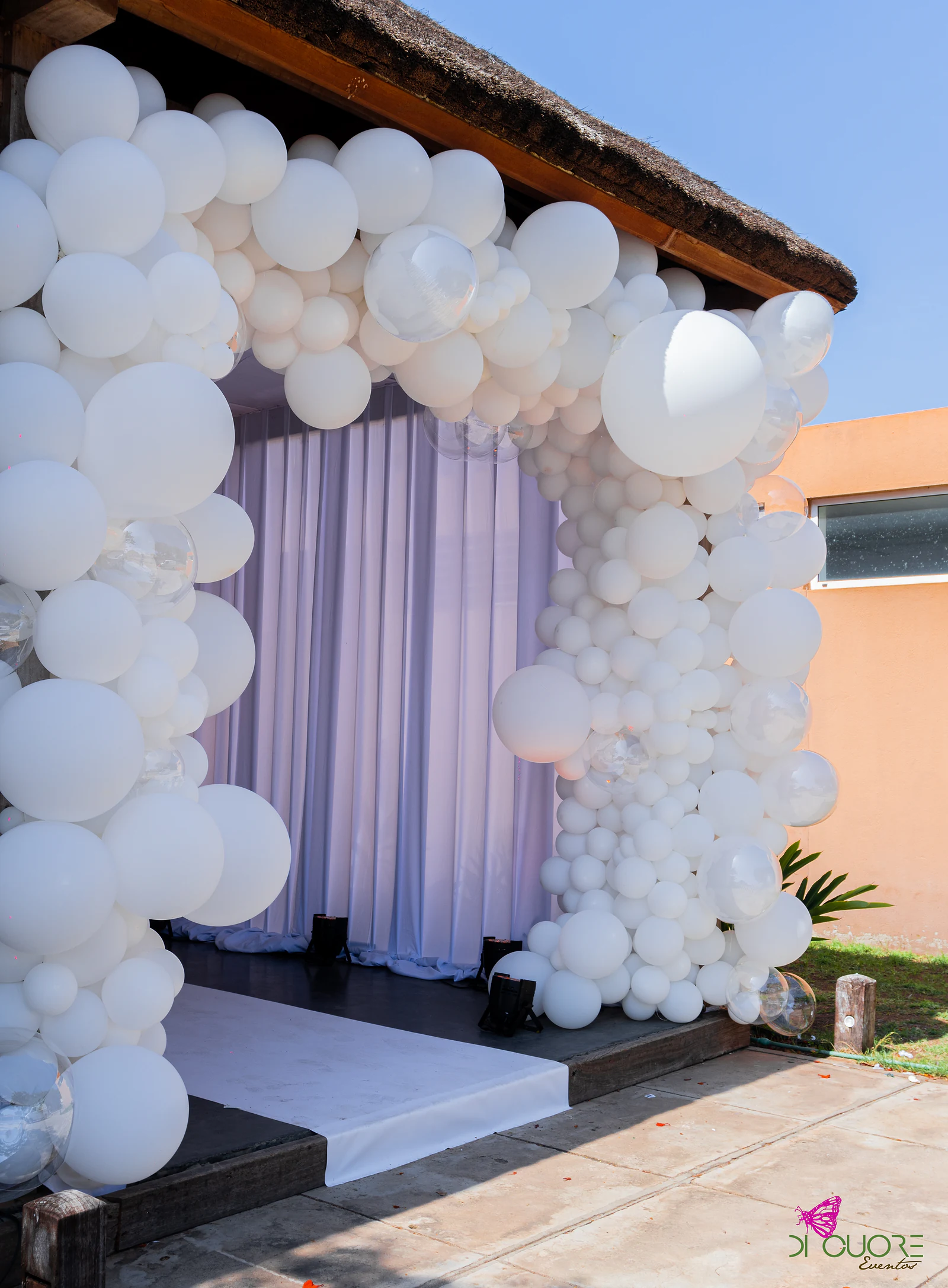 Arch of white balloons of assorted sizes around a curtain, beneath a thatched roof, with a white runner in front