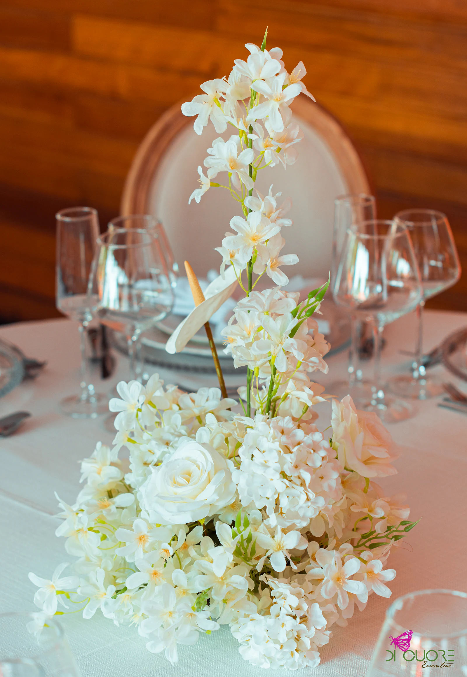 Tall centrepiece of white flowers, roses and anthurium, between crystal glasses and before a wooden wall