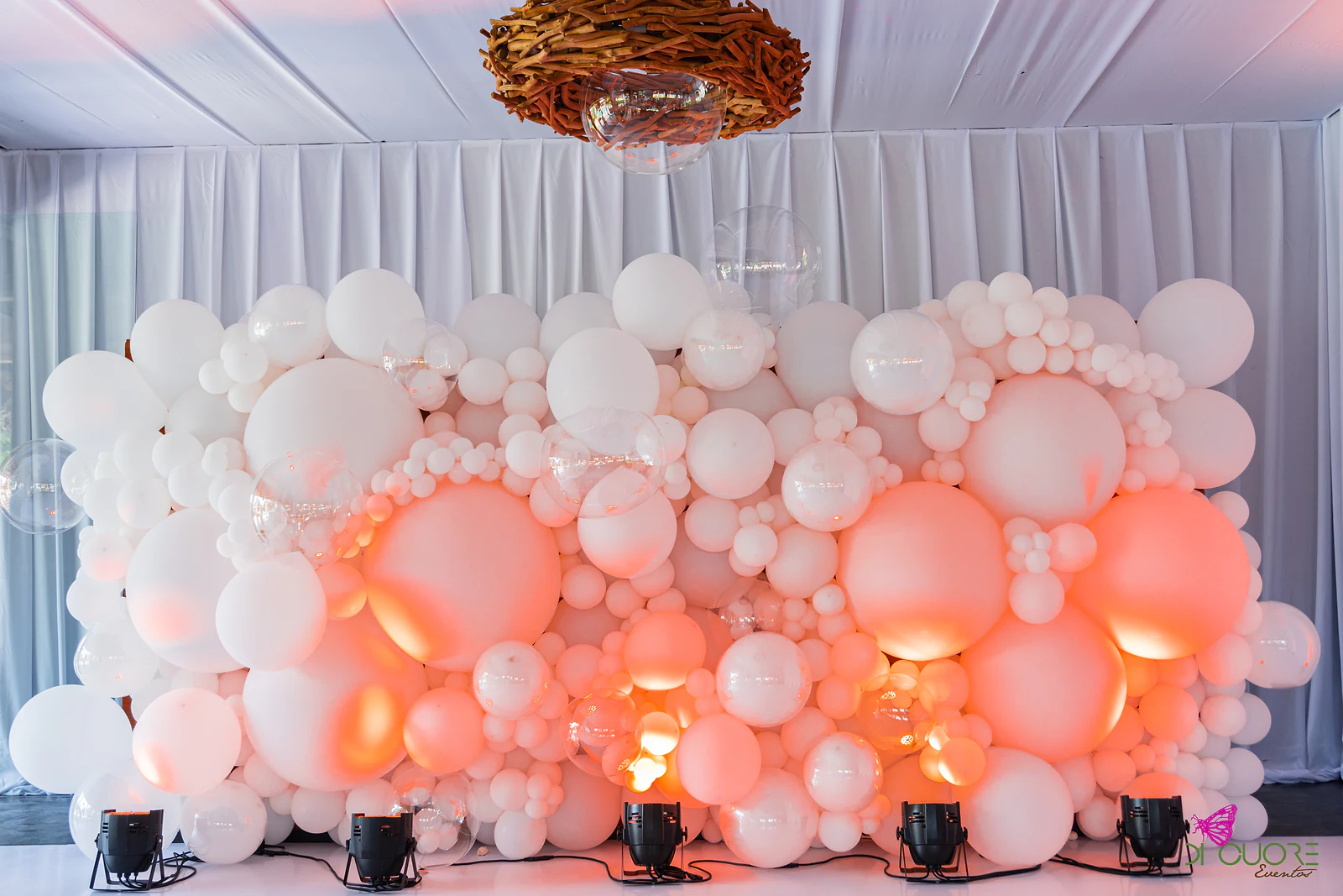 Wall of white balloons of assorted sizes lit in peach tones by spotlights, before a white curtain