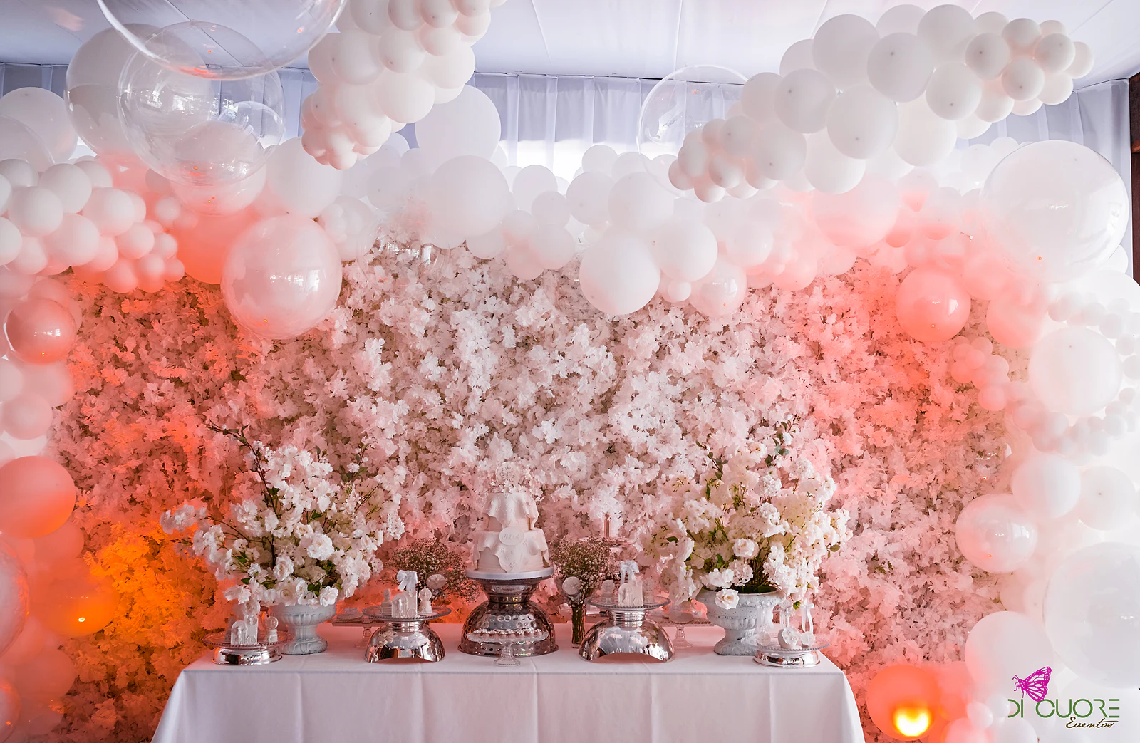Cake table before a wall of white and pale pink flowers, with an arch of white balloons above and silver stands holding sweets
