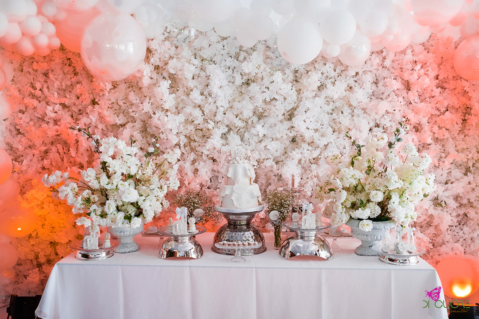 Mesa de doces de toalha branca com um bolo branco e baixelas de prata entre jarras de flores brancas, diante de uma parede de flores brancas e cor-de-rosa com balões brancos