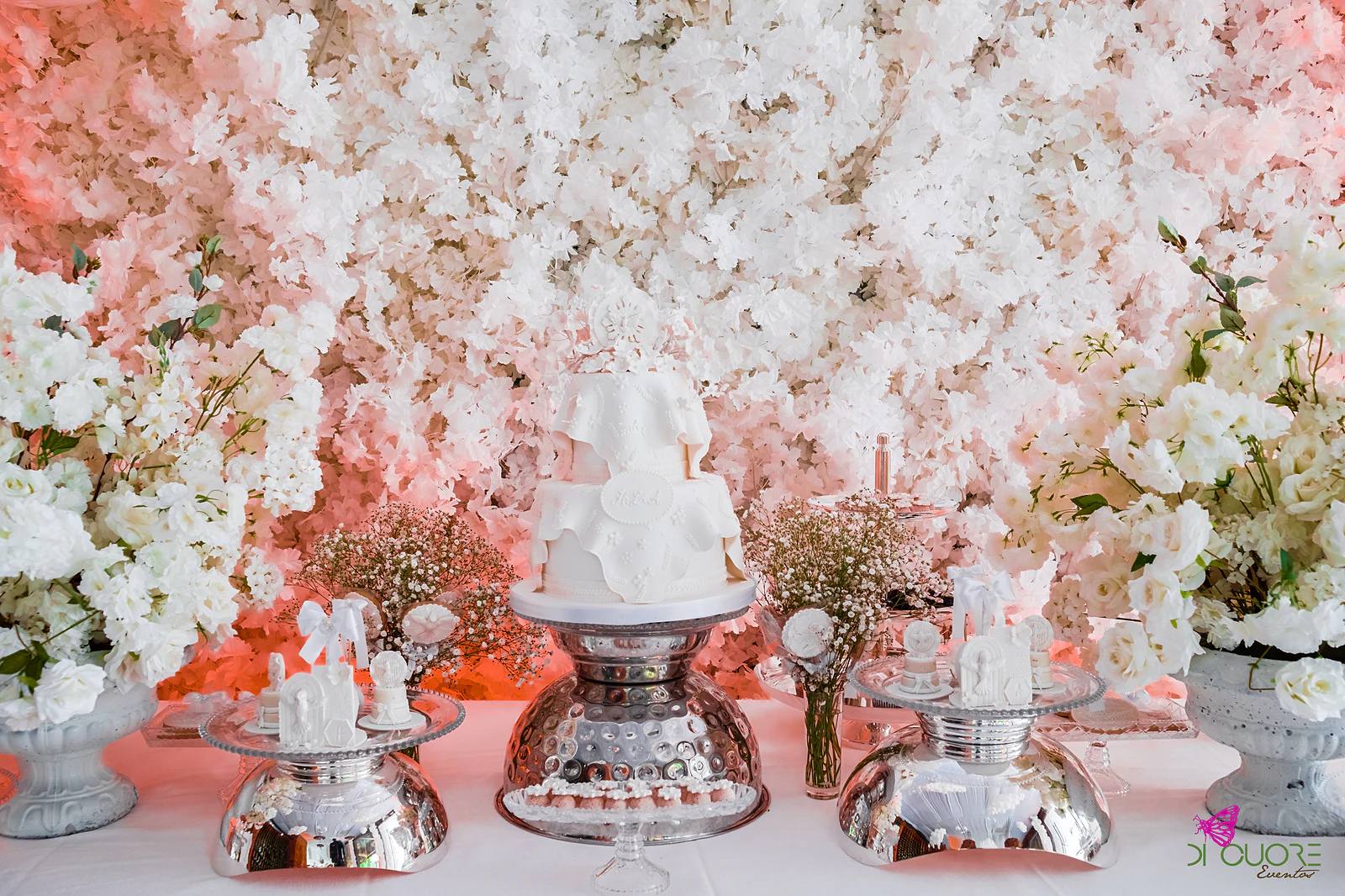 White cake and sweets on mirrored stands, between urns of white flowers, before a flower wall