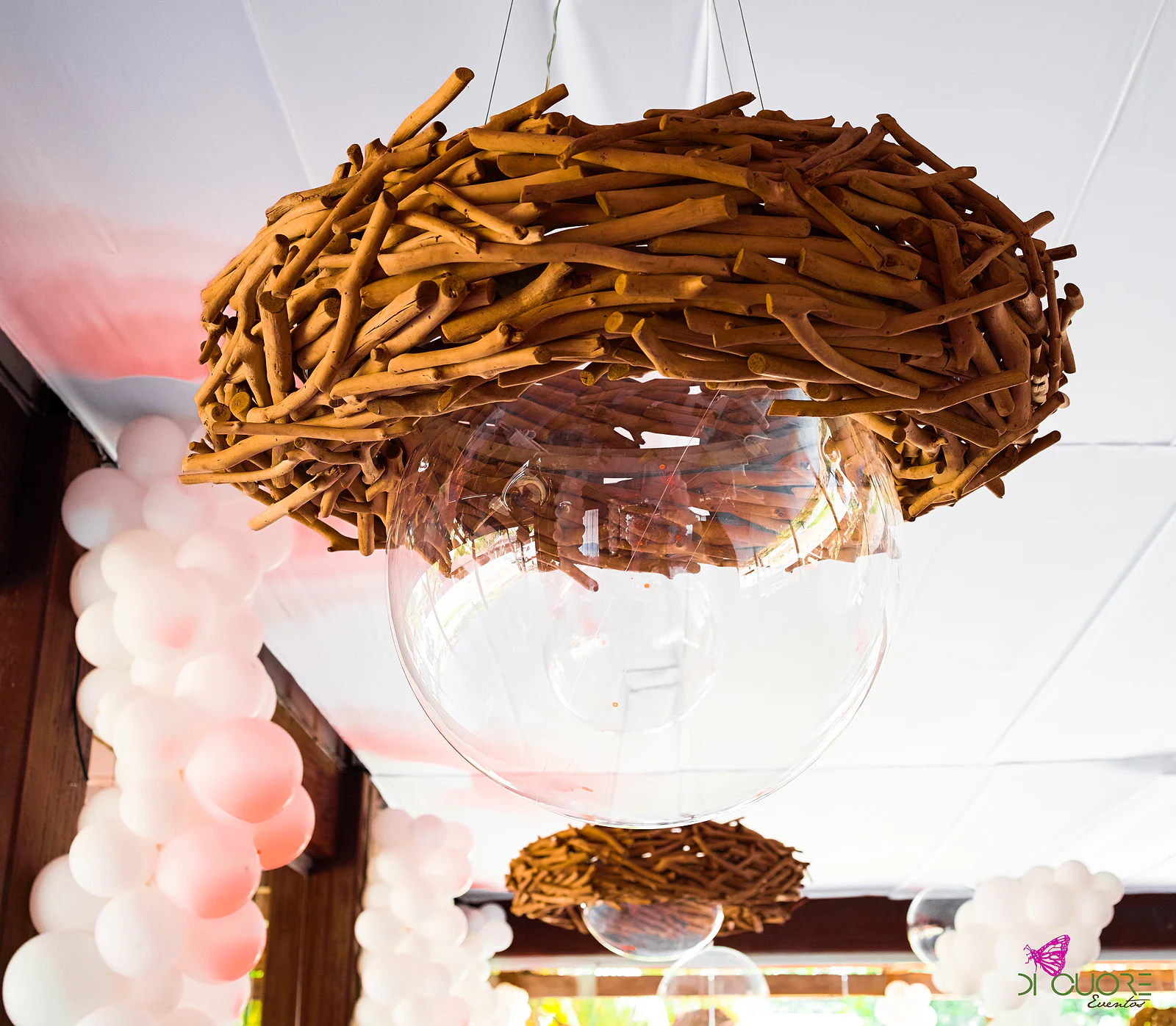 Suspended twig nest with a glass globe, beside a column of white and pale pink balloons