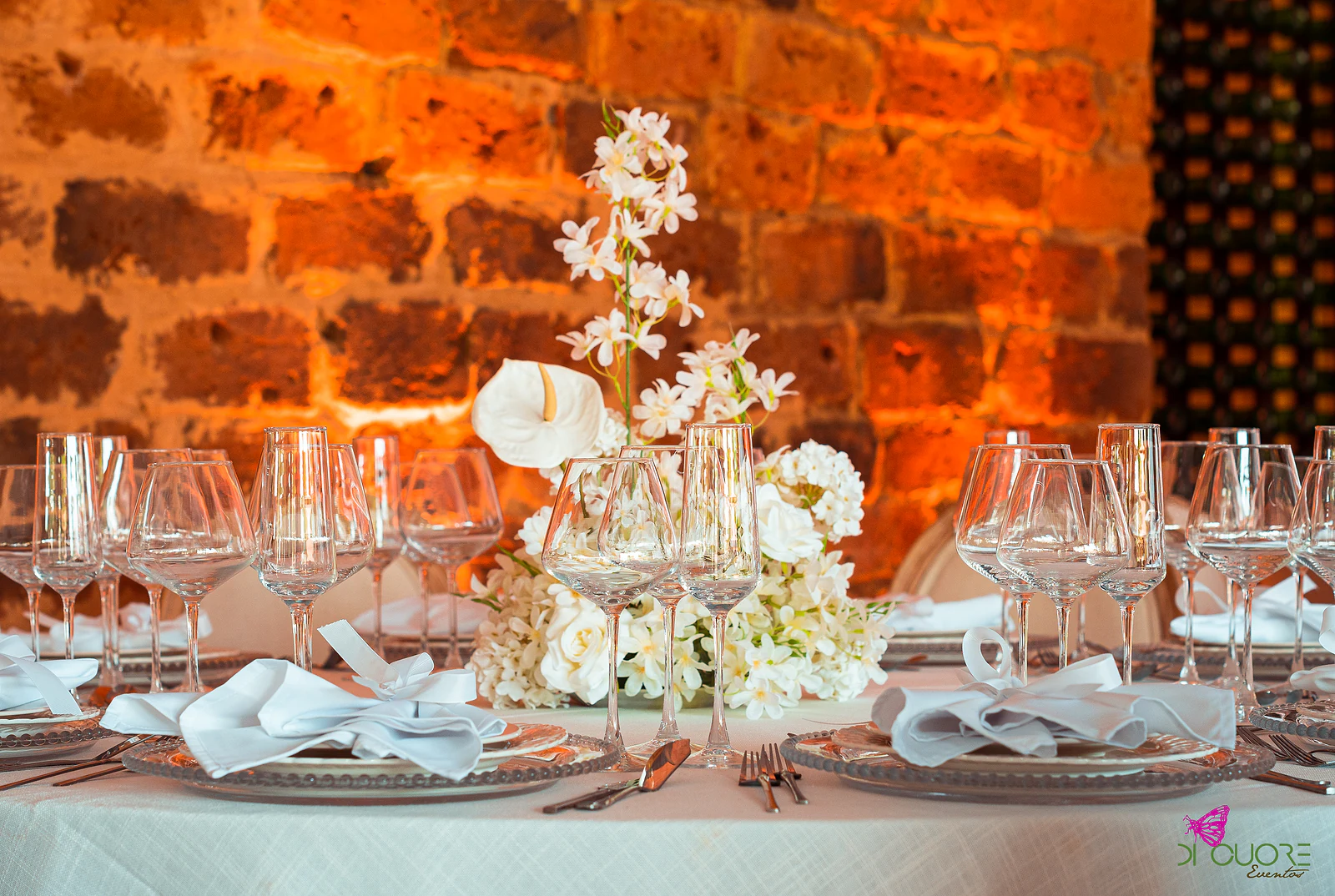 Laid table before an orange-lit brick wall, with a tall spray of white flowers, anthurium and ribbon-tied napkins