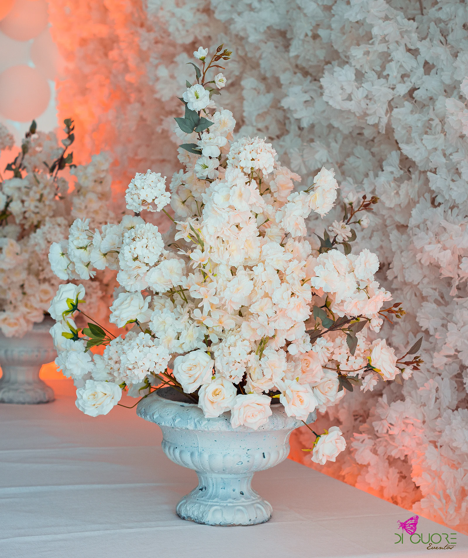 Weathered white urn holding a spray of white flowers and roses, before a flower wall in peach-coloured light