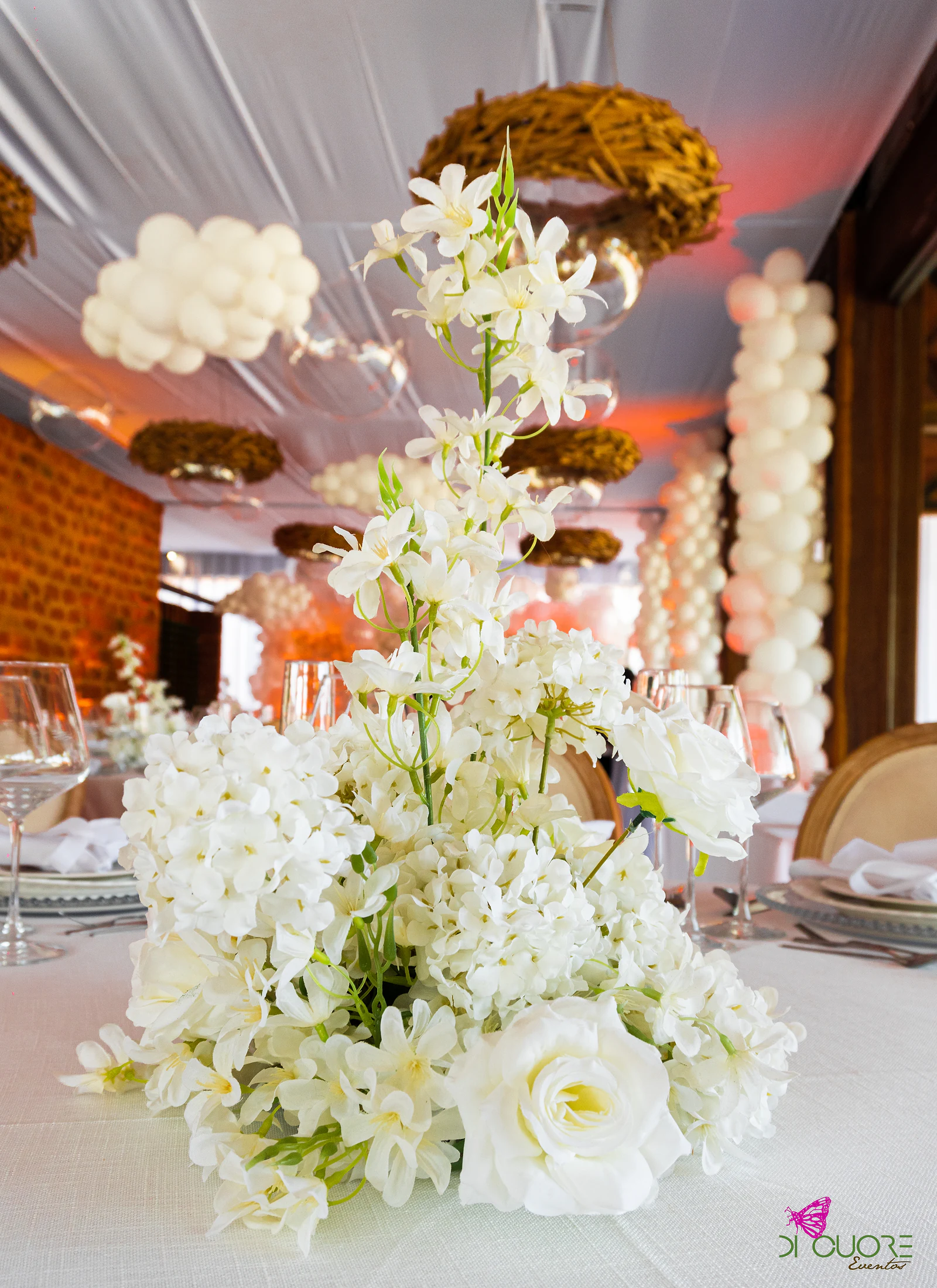 Tall centrepiece of white flowers and roses on a pale cloth, with balloon columns and hanging nests behind