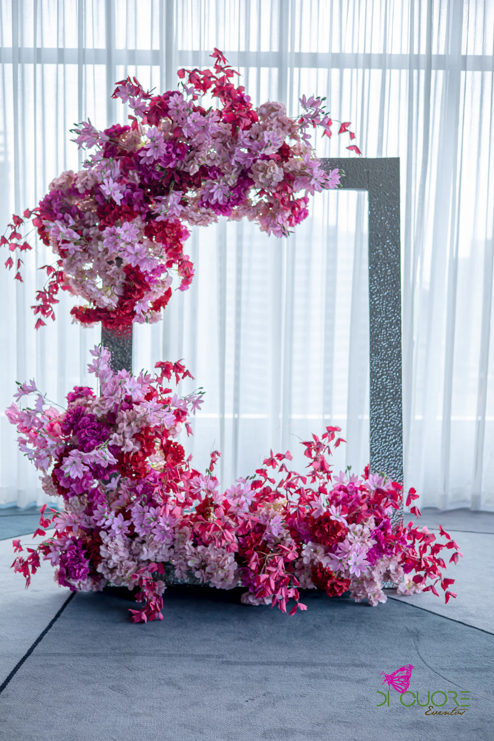 Crescent-shaped installation of pink and fuchsia flowers with a silver frame and white curtains behind