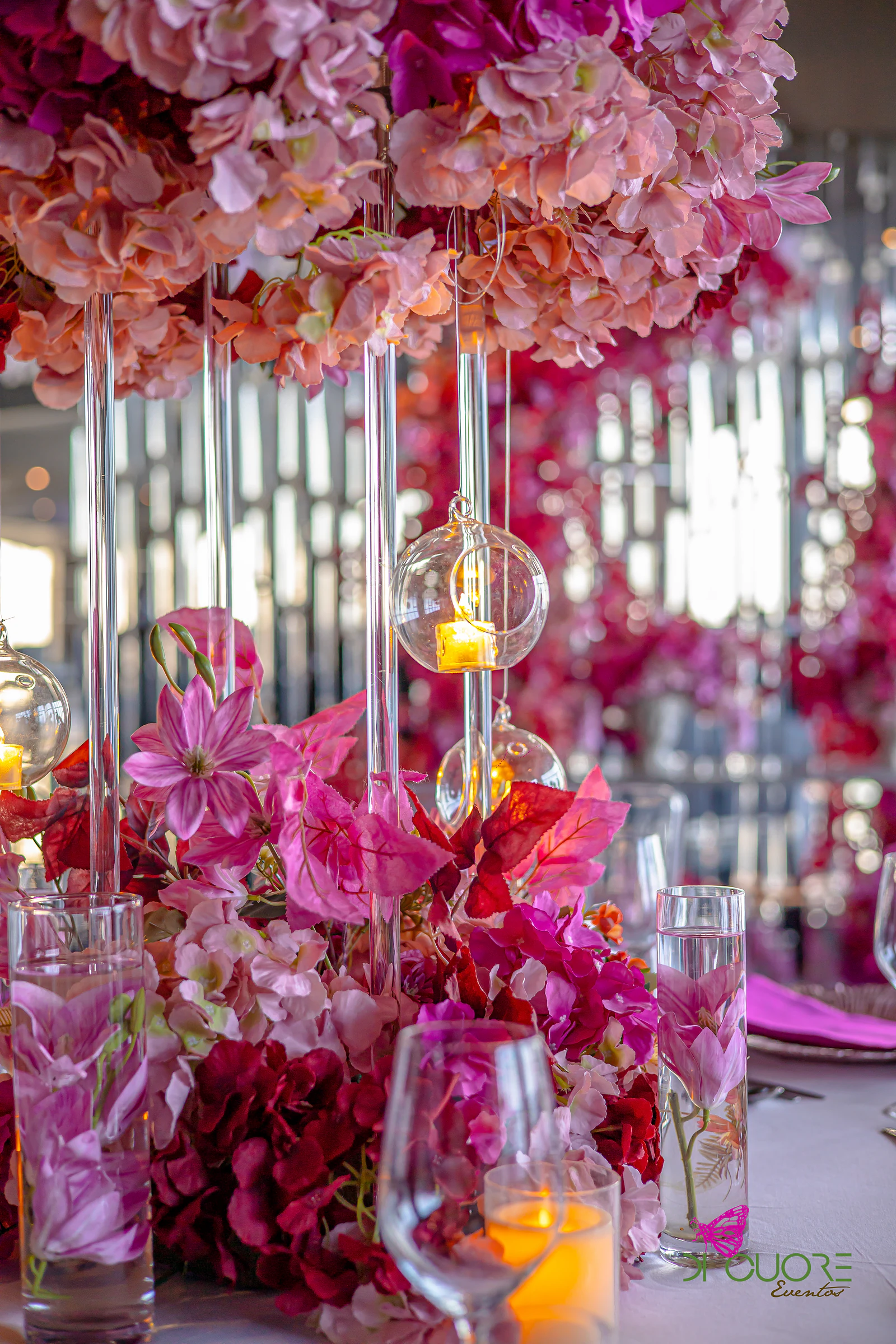 Tall glass stems with hanging lanterns and candles, beneath a globe of pink and peach flowers, above an arrangement of fuchsia and red blooms