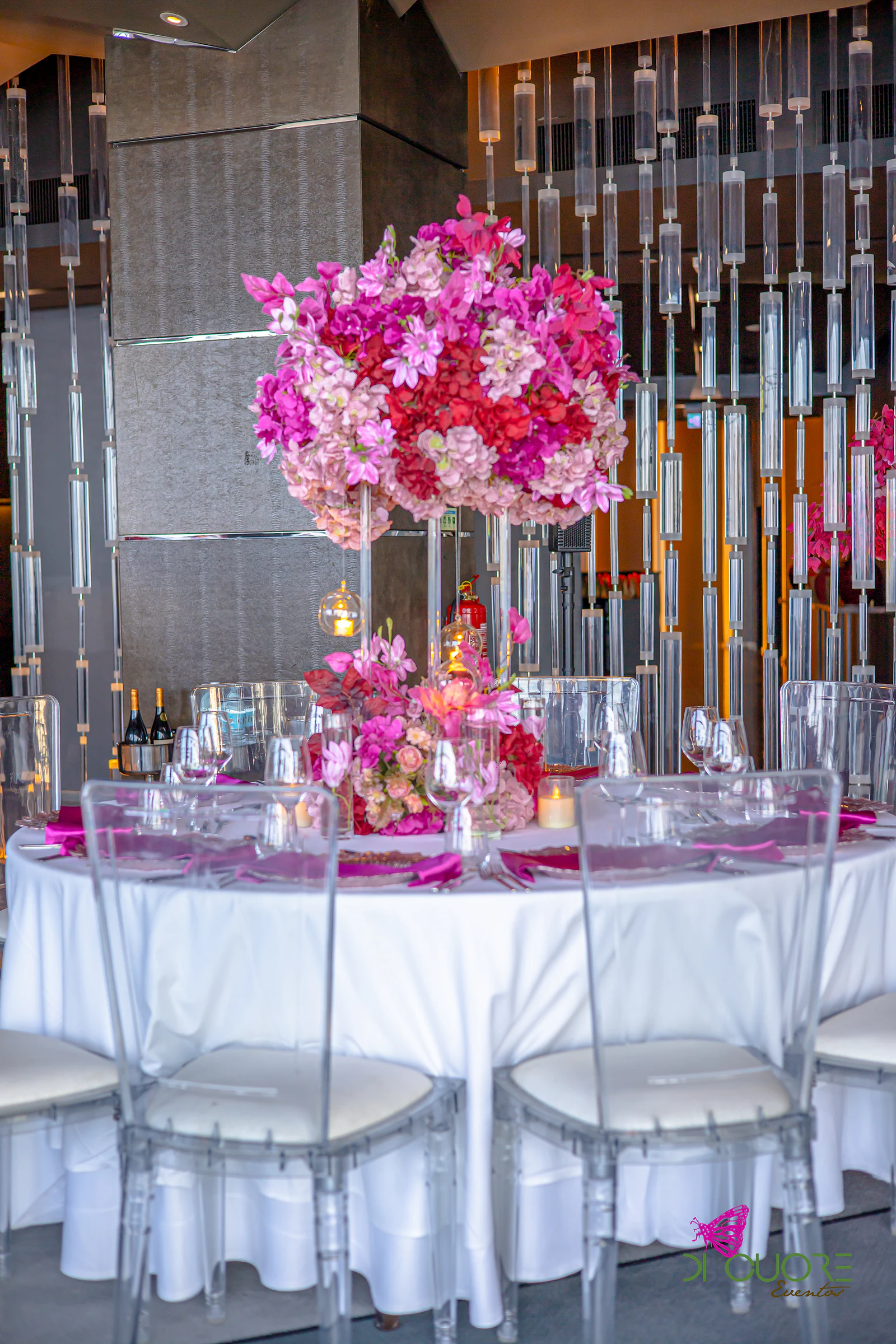Round table in white linen with acrylic chairs and a tall pink and red floral centrepiece, before the crystal wall