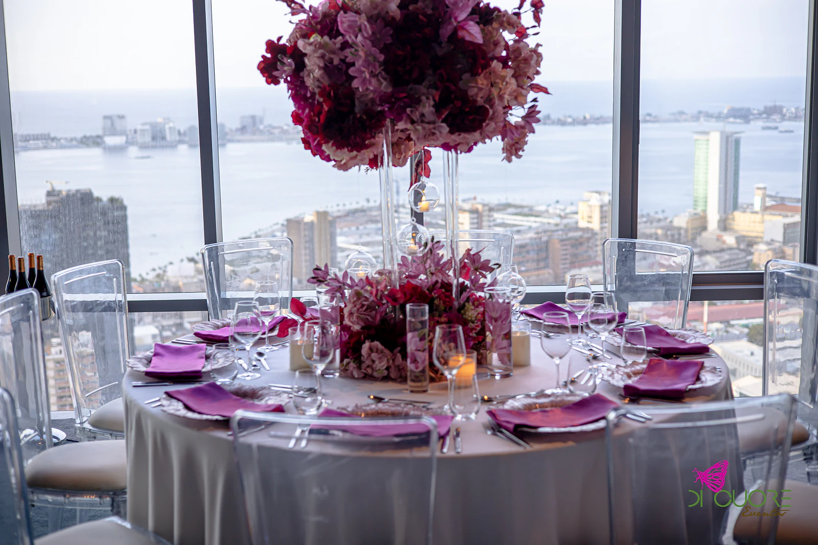 Round table with a tall burgundy floral centrepiece and fuchsia napkins, before a panoramic window over the city and the sea