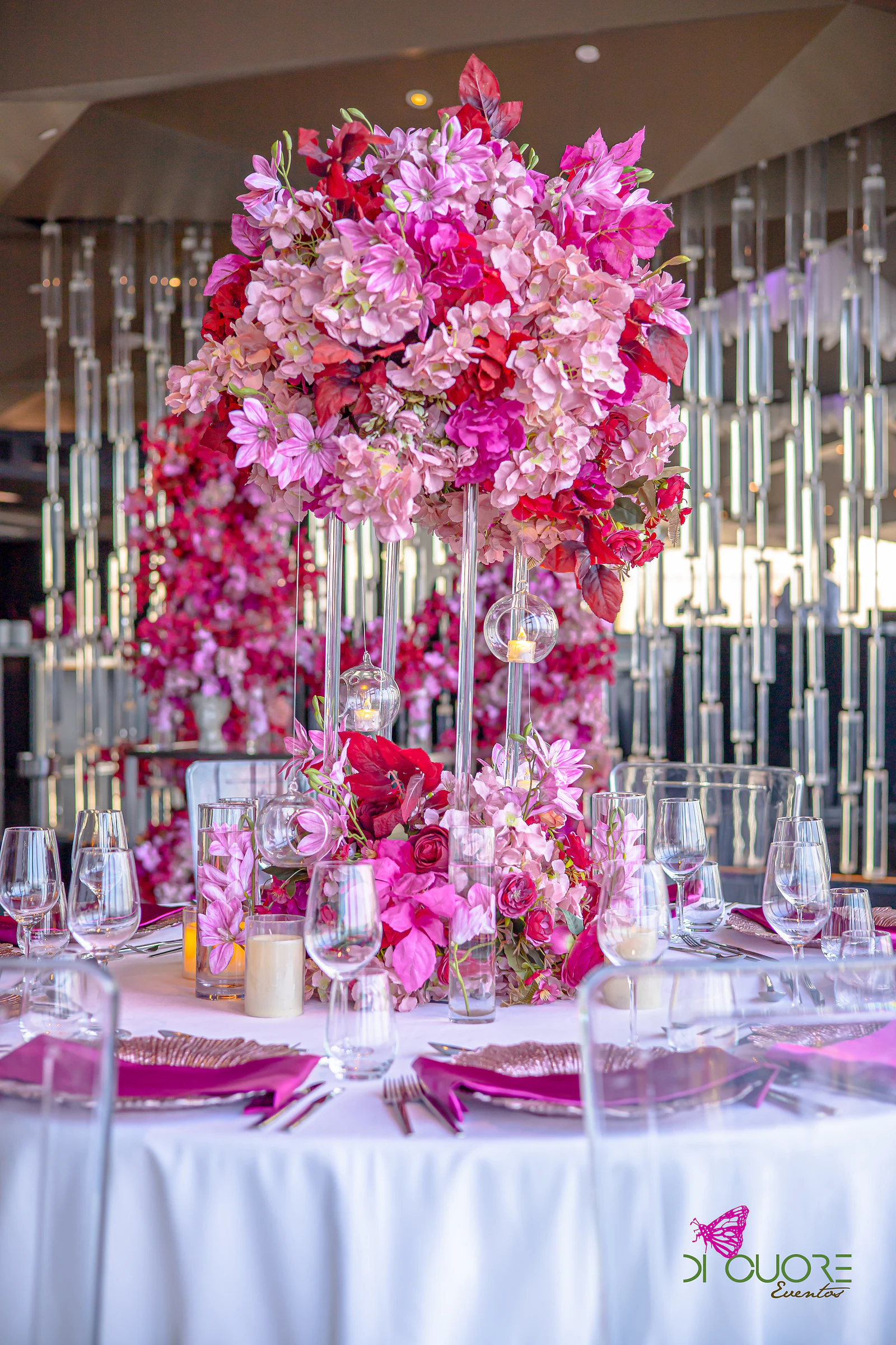 Round table with a tall pink and red floral centrepiece on glass stems, with hanging lanterns and the crystal wall behind