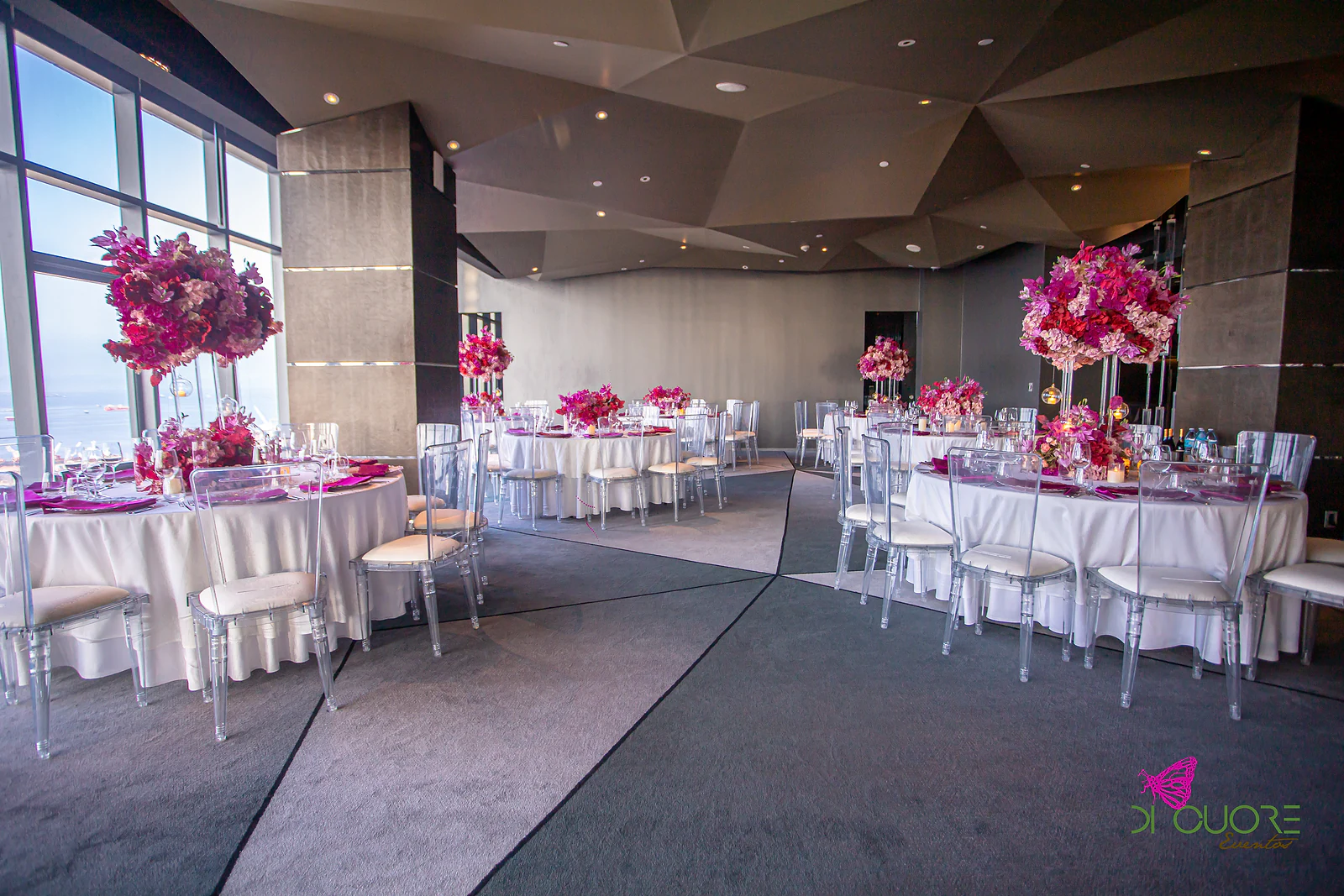 Spacious room with a faceted ceiling, round tables in white linen, acrylic chairs and tall pink floral centrepieces, with windows over the sea