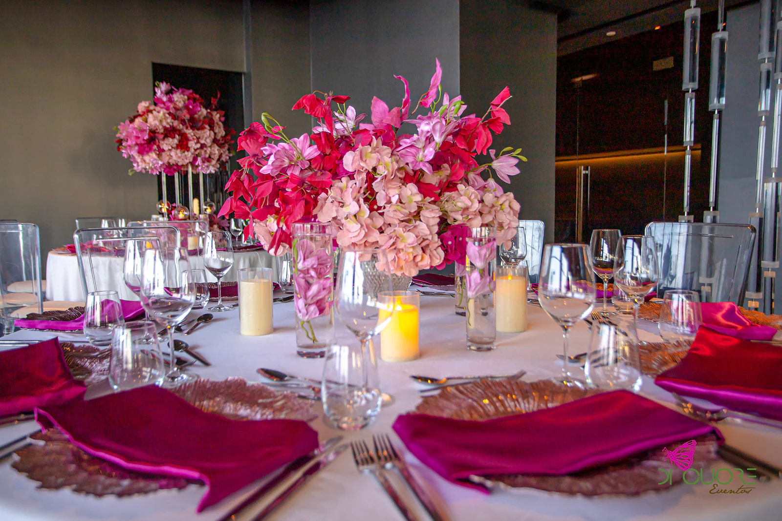 Laid table with deep fuchsia satin napkins on copper chargers, candles and a tall arrangement of pink and red flowers
