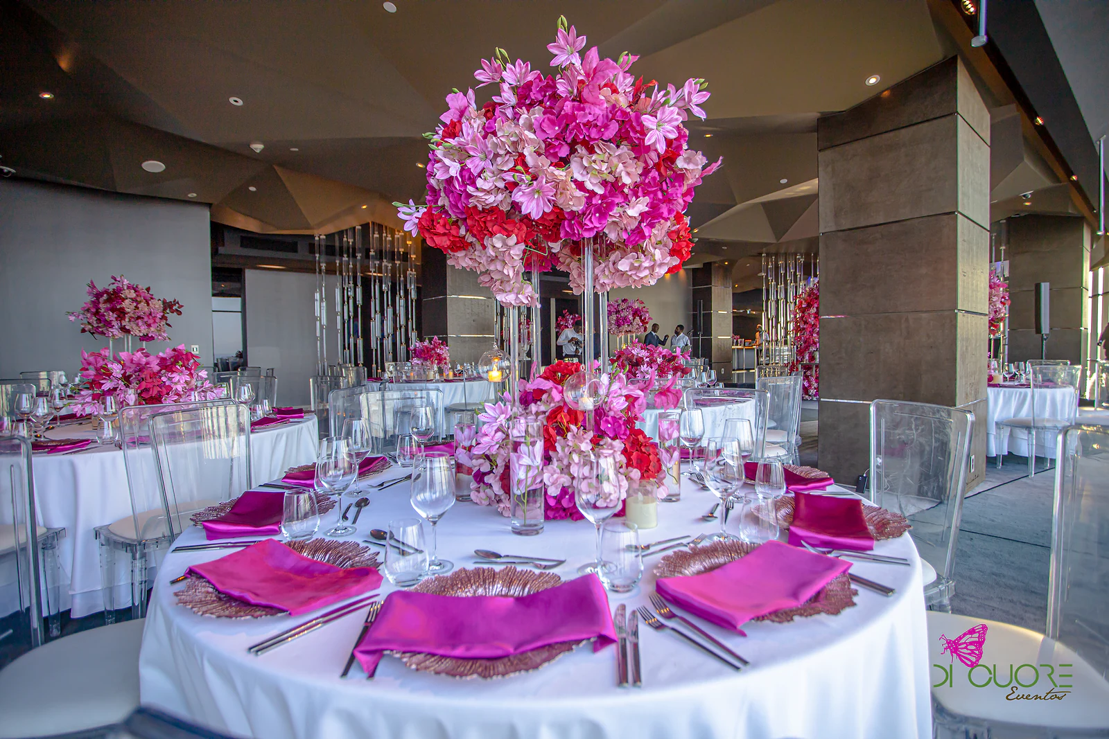 Round table in white linen with fuchsia napkins on copper chargers and a tall pink and red floral centrepiece, in a room with a faceted ceiling
