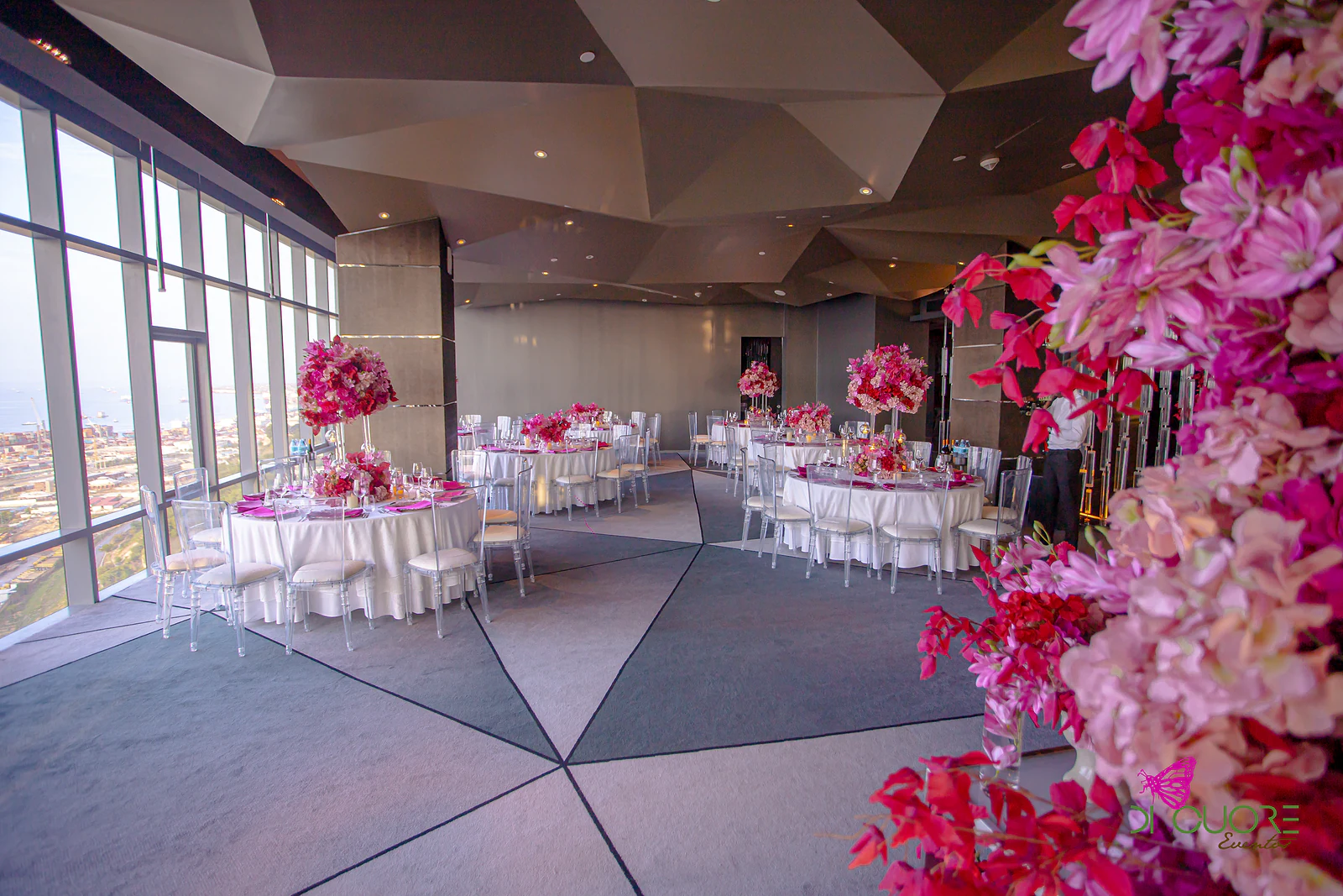 Room with a faceted ceiling, panoramic windows and laid round tables, with pink and red flowers in the foreground