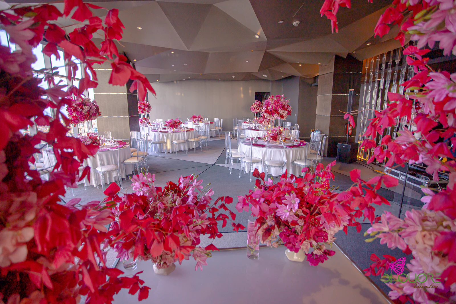 The room seen through red and pink flowers in the foreground, with round tables and a faceted ceiling beyond