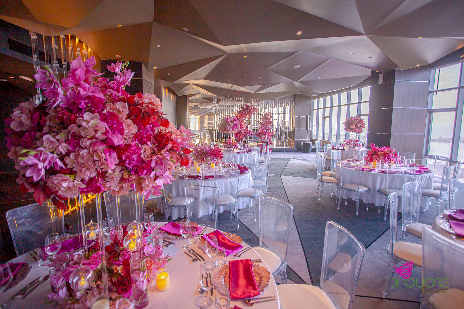 Tall pink and fuchsia floral centrepiece in the foreground and rows of round tables beyond, under warm light and a faceted ceiling