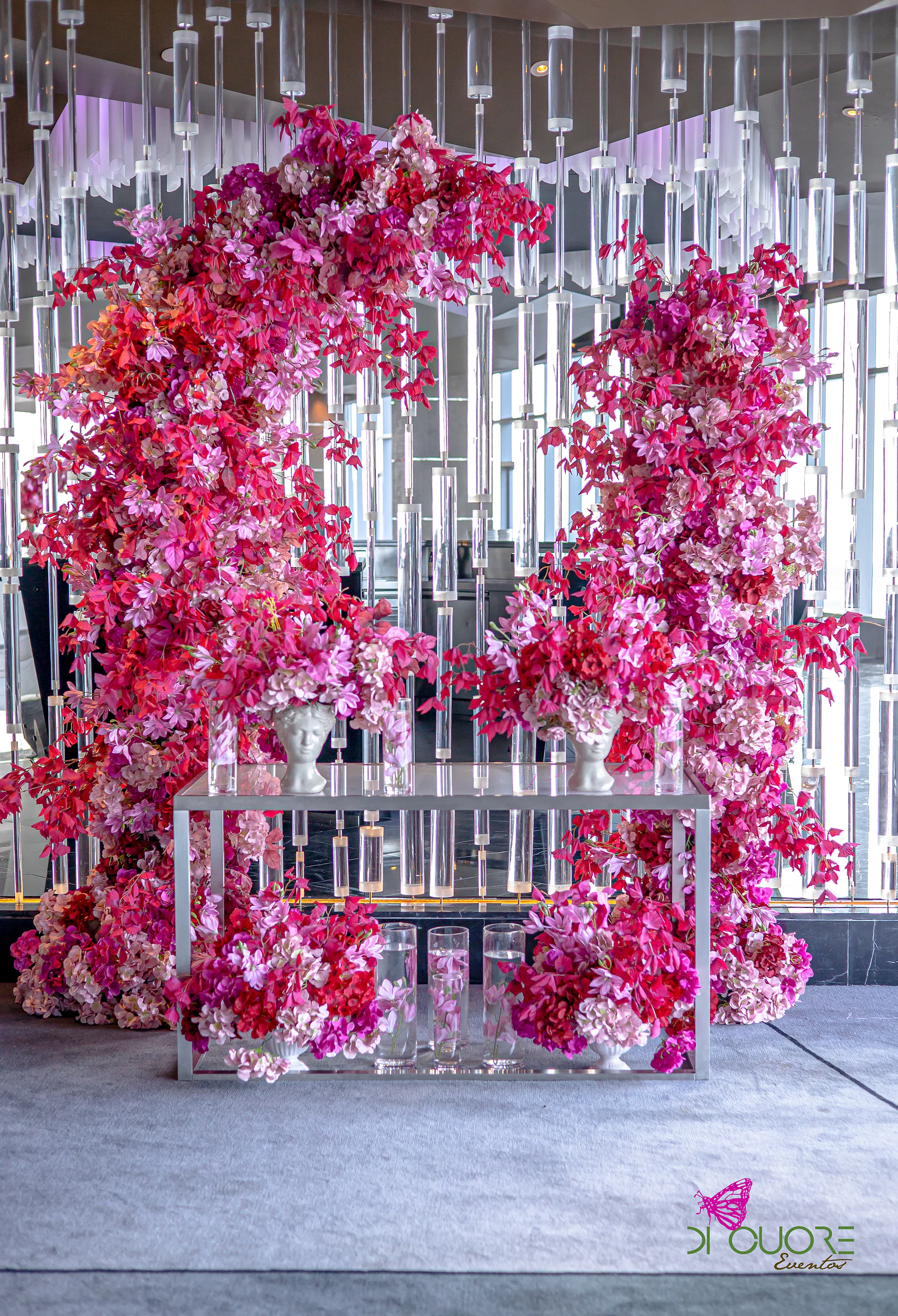 Dois arcos de flores fúcsia, rosa e vermelho diante de uma parede de hastes de cristal, com uma consola prateada e vasos brancos em forma de busto