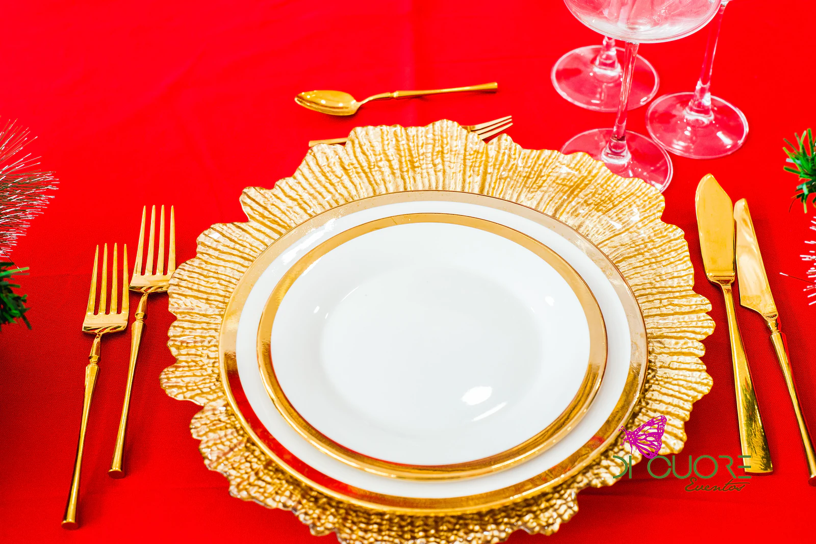Place setting with a white plate on a gold charger, gold cutlery and stemmed glasses, on a red cloth