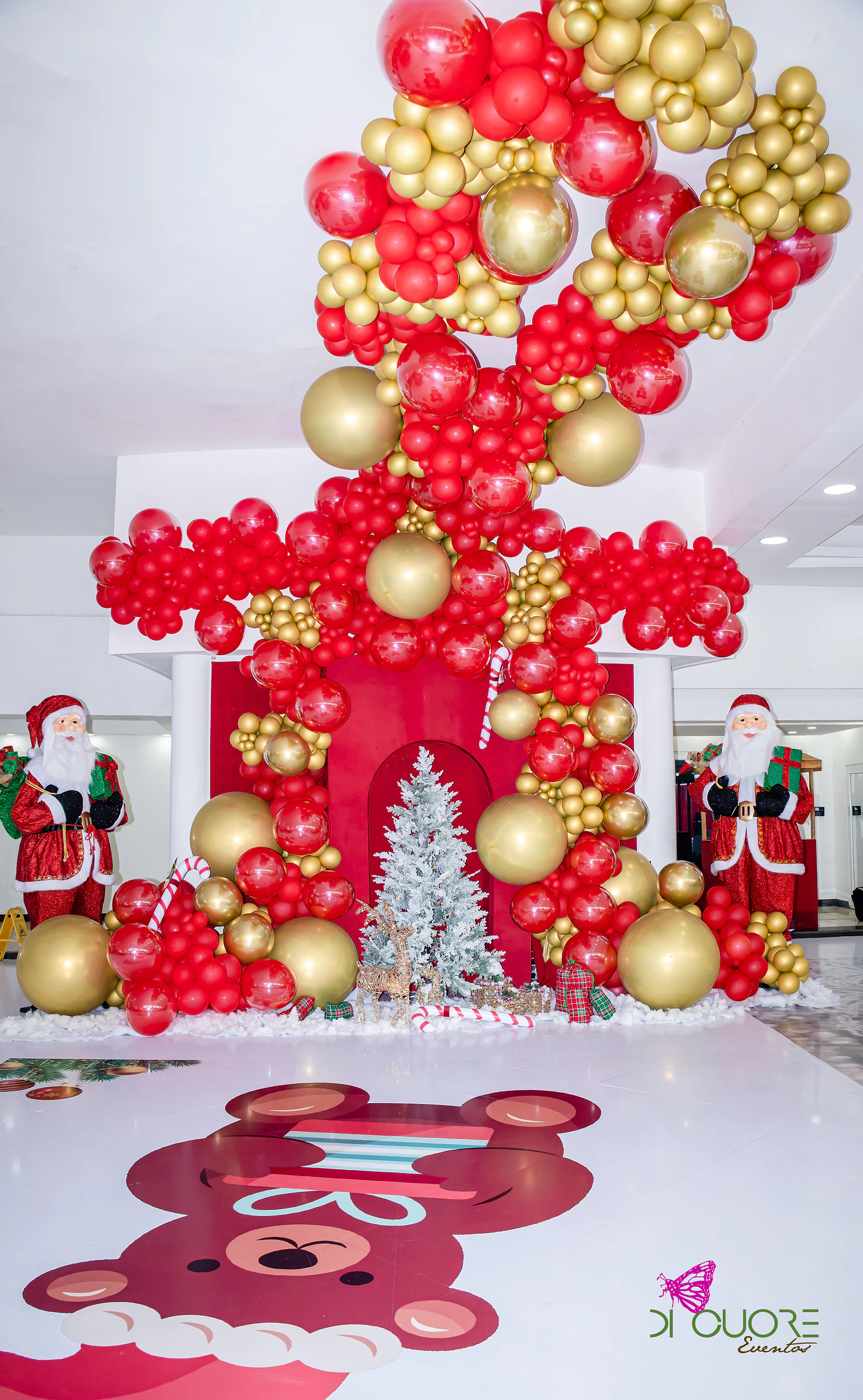 Escultura de balões vermelhos e dourados sobre uma lareira vermelha, com árvore prateada e duas figuras do Pai Natal, e um urso de Natal desenhado no chão branco