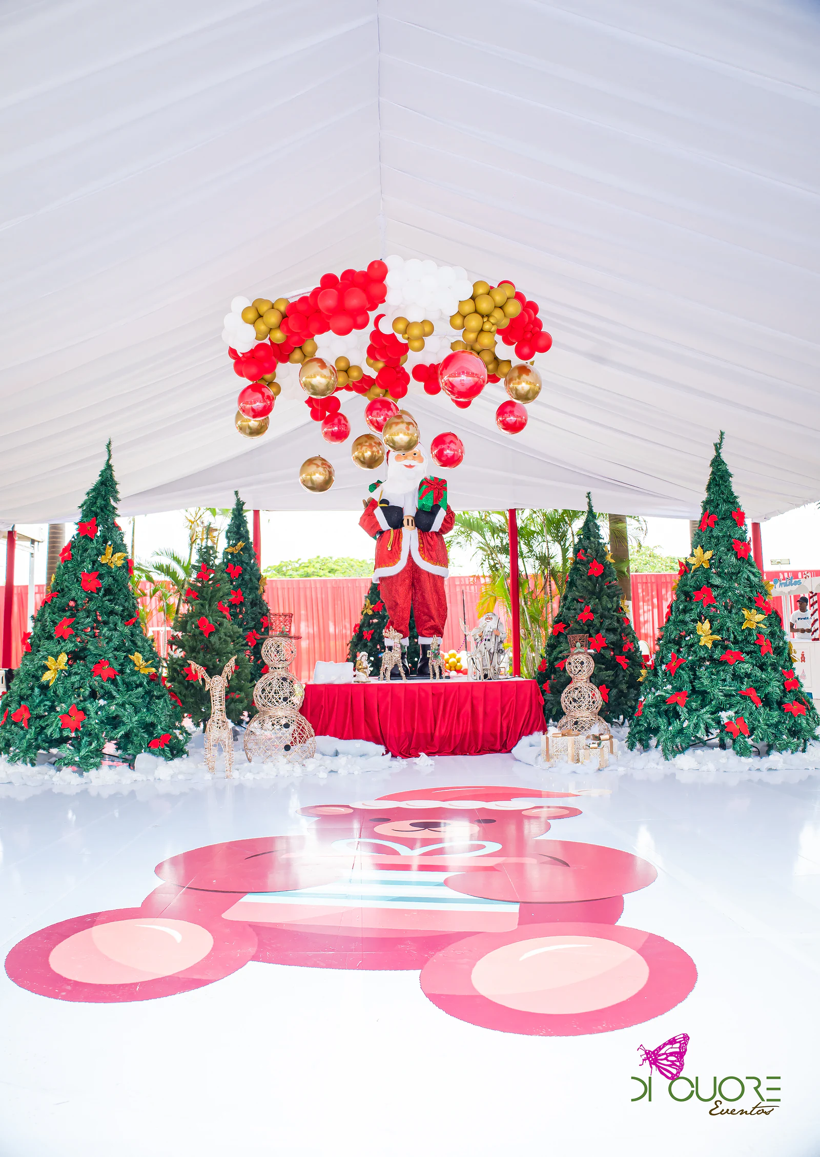 White tent with Father Christmas among Christmas trees, beneath a chandelier of red and gold balloons, and a Christmas bear drawn on the white floor