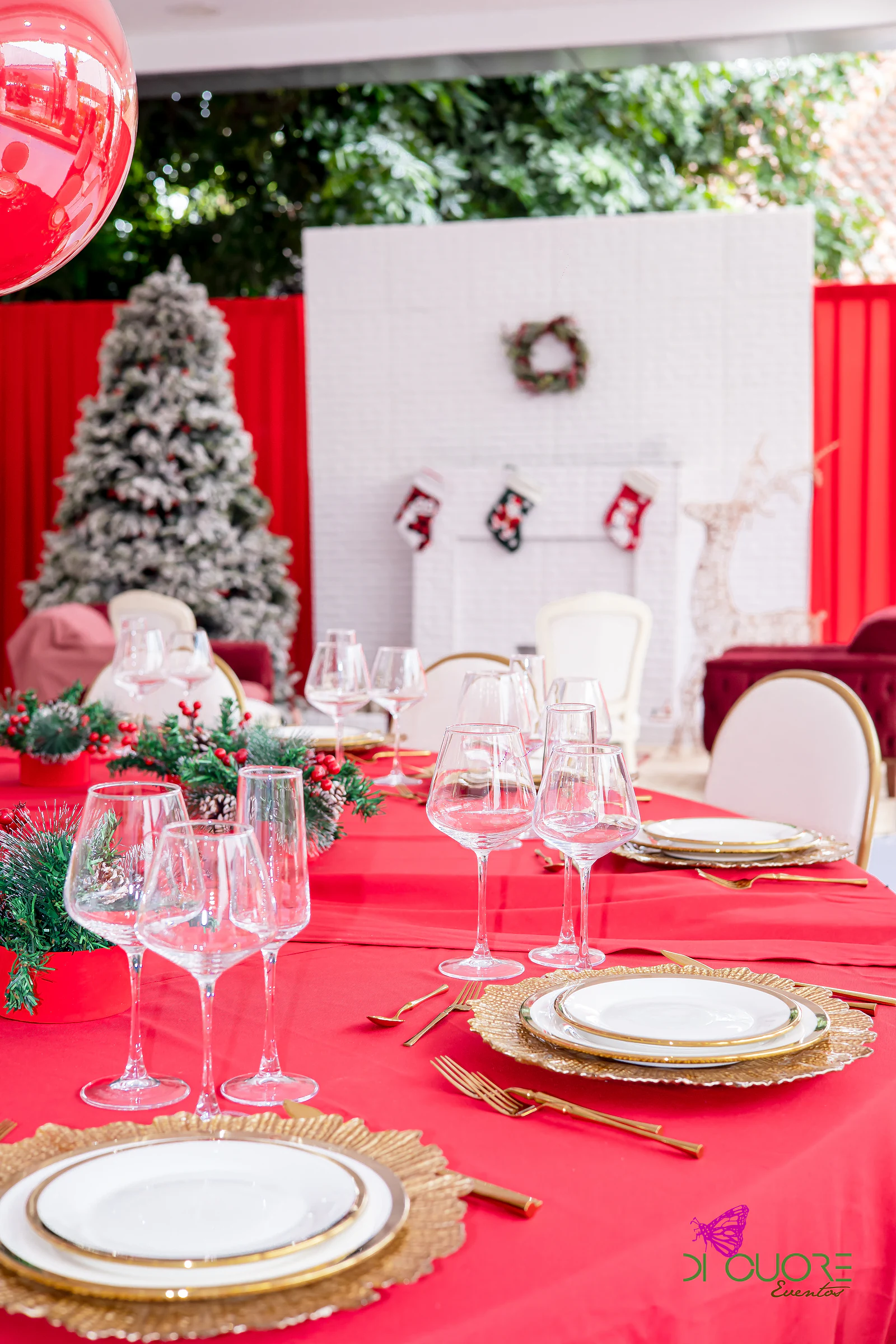 Red-clothed table with gold chargers, crystal glasses and pine arrangements, the white fireplace and snow-dusted tree behind