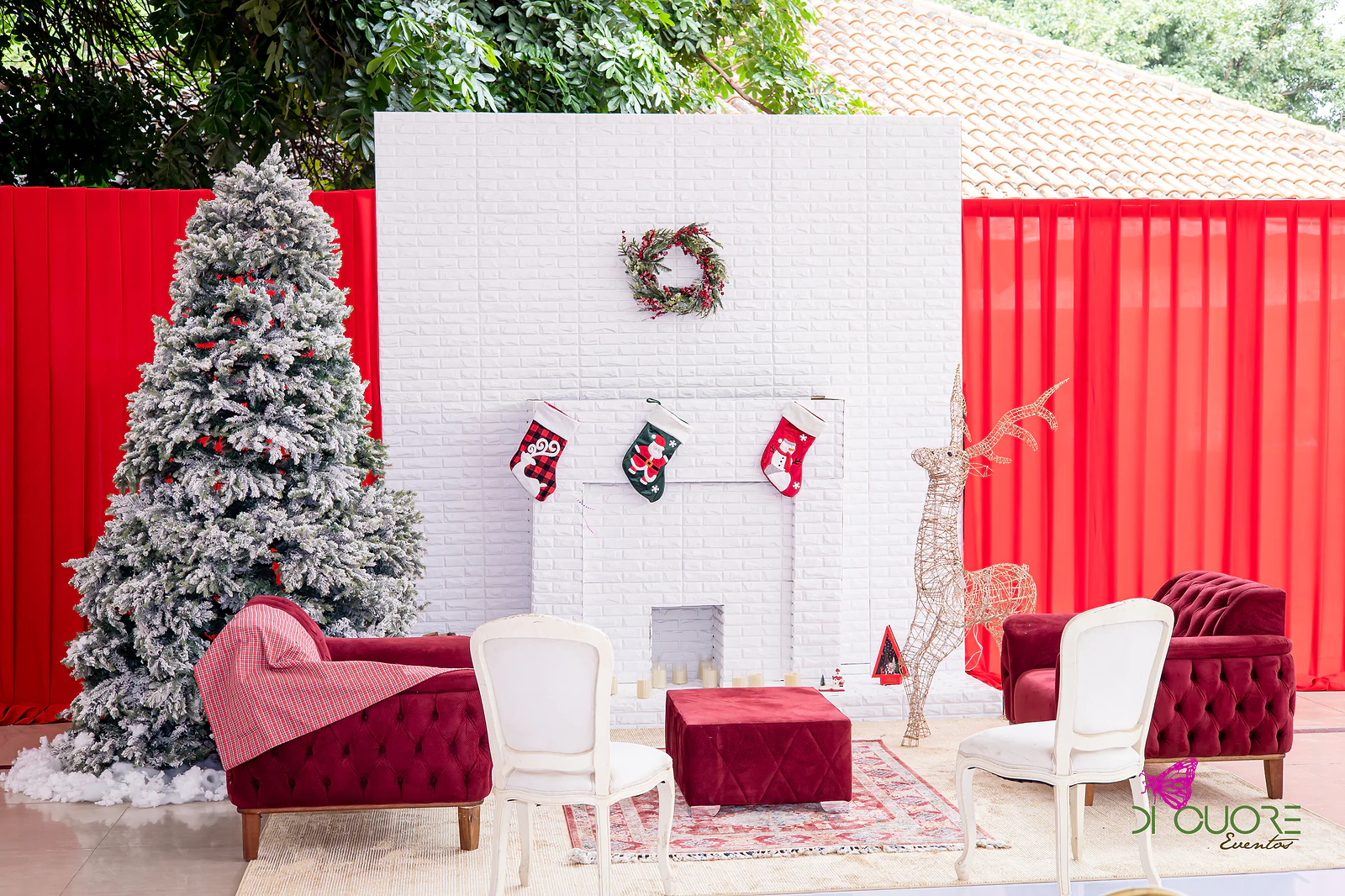 Corner with a white fireplace, Christmas stockings and wreath, a snow-dusted tree, a wire reindeer and burgundy velvet armchairs on a rug, before red drapes