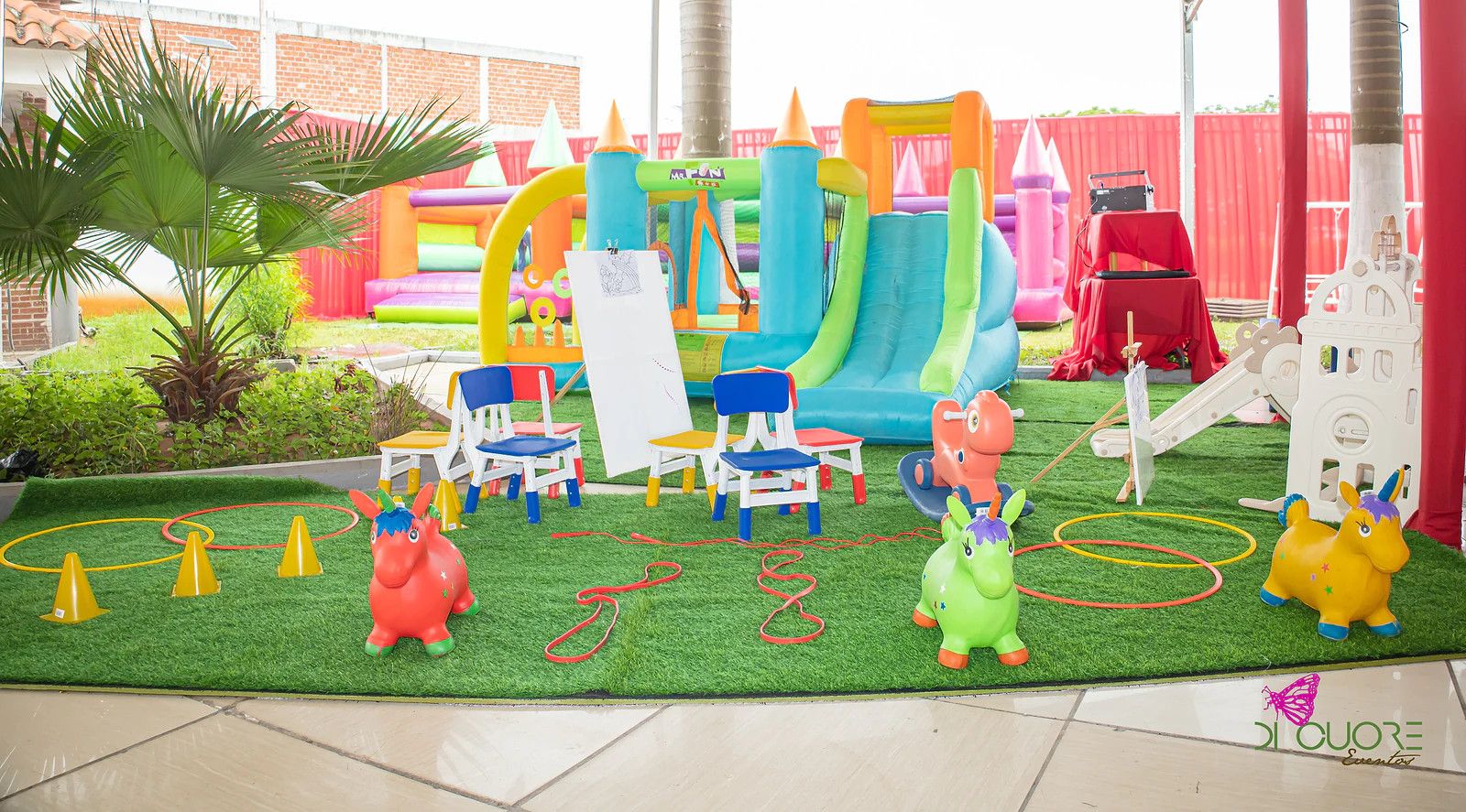 Grassy corner with colourful bouncy castles, hopping horses, little chairs, painting easels, hoops and cones