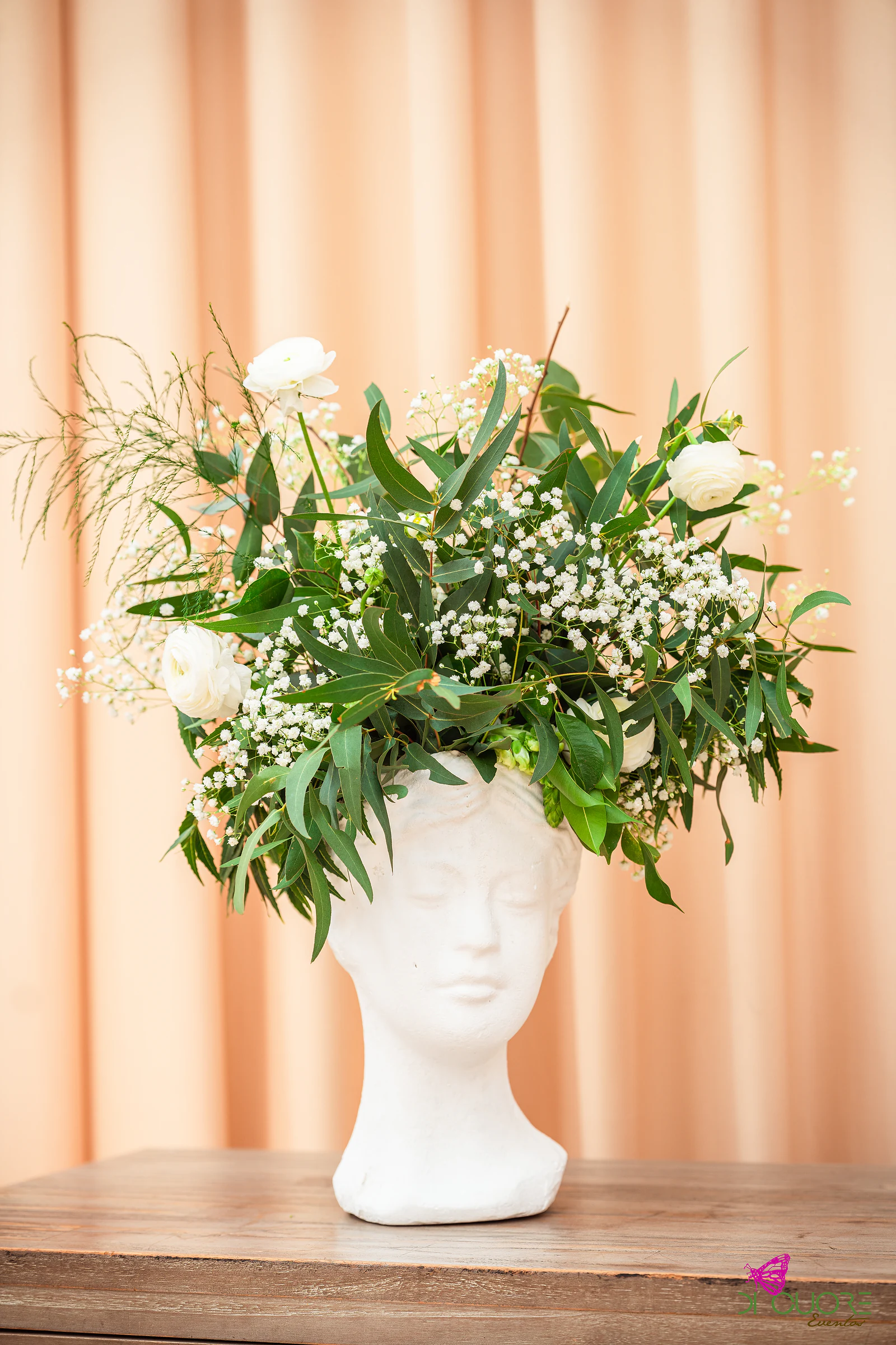Vaso branco em forma de busto com ramo de flores brancas e folhagem, sobre um aparador de madeira