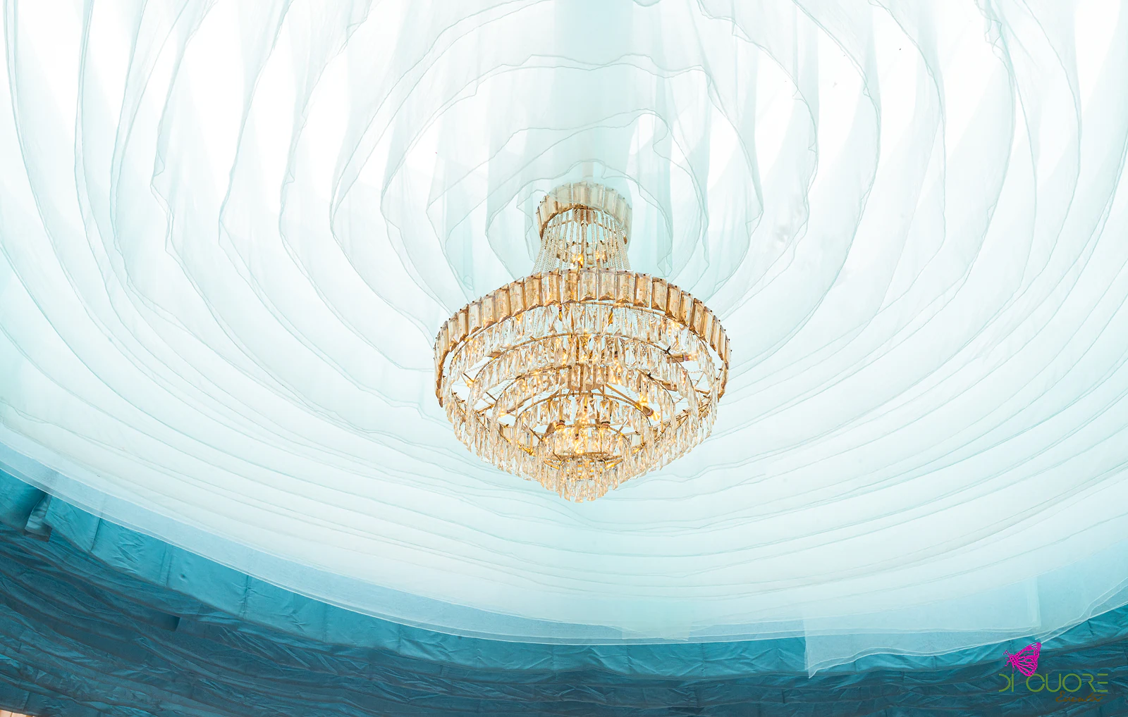 Lustre de cristal dourado sob o tecto de tecido branco e azul-turquesa, com cortinados cor de pêssego a toda a volta