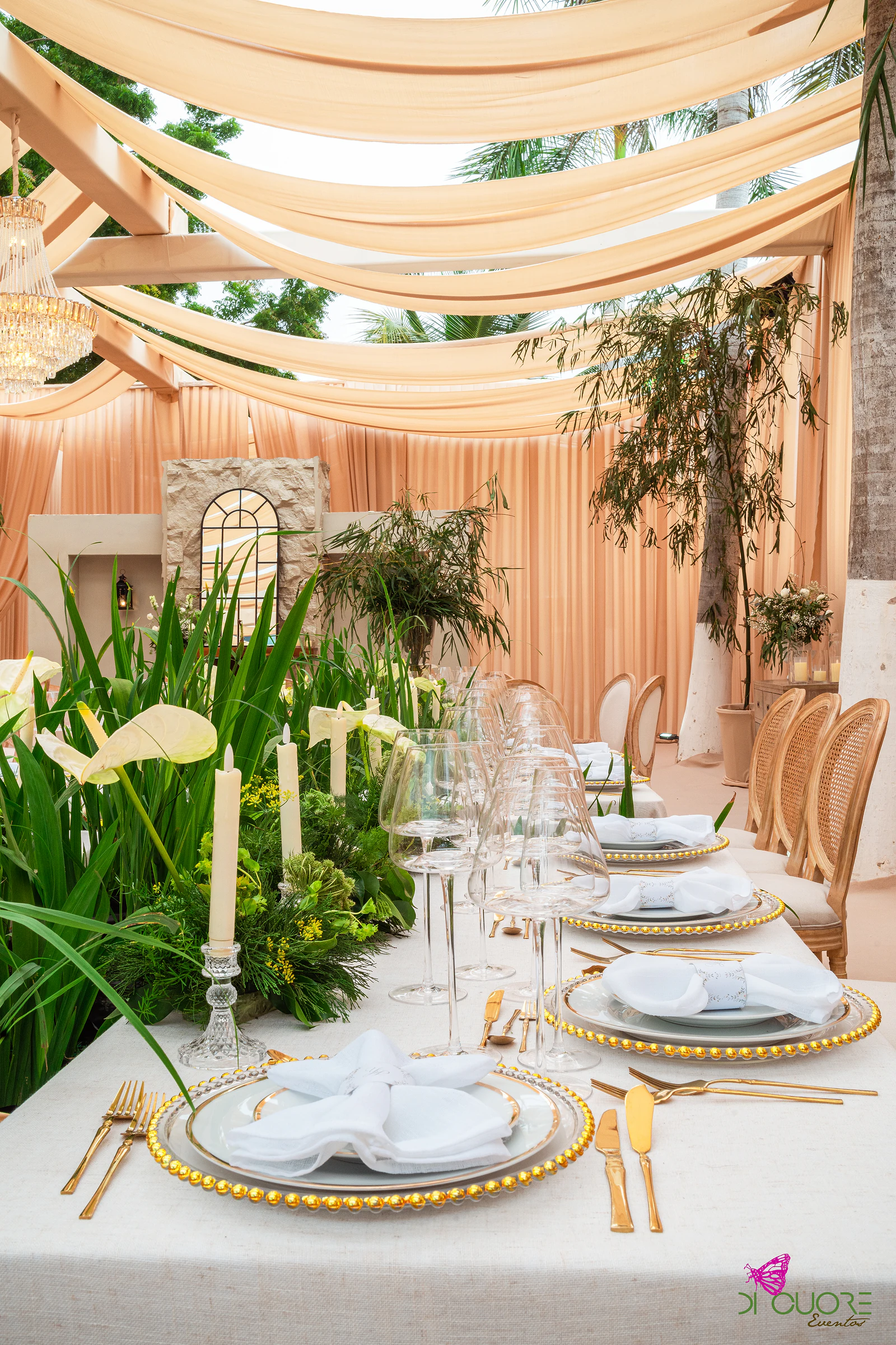 Mesa posta com velas altas, folhagem e flores brancas, pratos com contas douradas e tecidos cor de pêssego no tecto