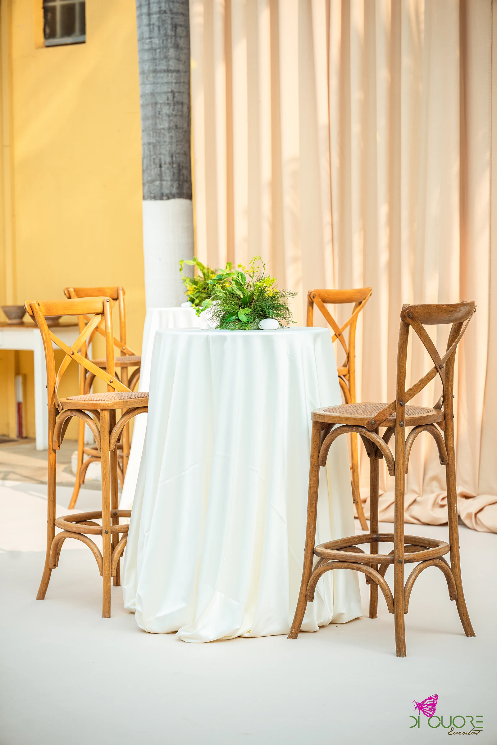 Mesa alta com toalha branca e uma pequena composição de folhagem, entre bancos altos de madeira, junto a cortinados cor de pêssego