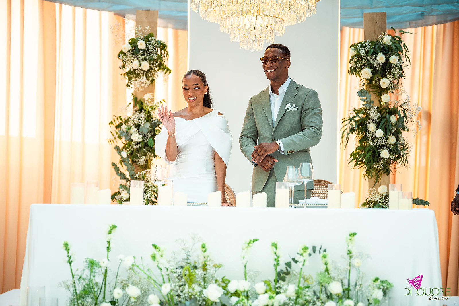 Os noivos de pé atrás da mesa dos noivos, ela de branco e ele de verde-claro, a sorrir entre colunas de folhagem e flores brancas