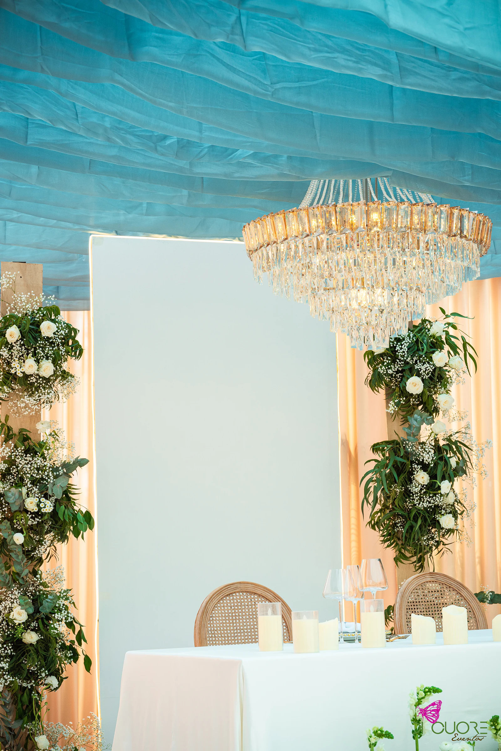 Painel branco entre colunas de folhagem e rosas brancas, com um lustre de cristal e a mesa dos noivos com velas